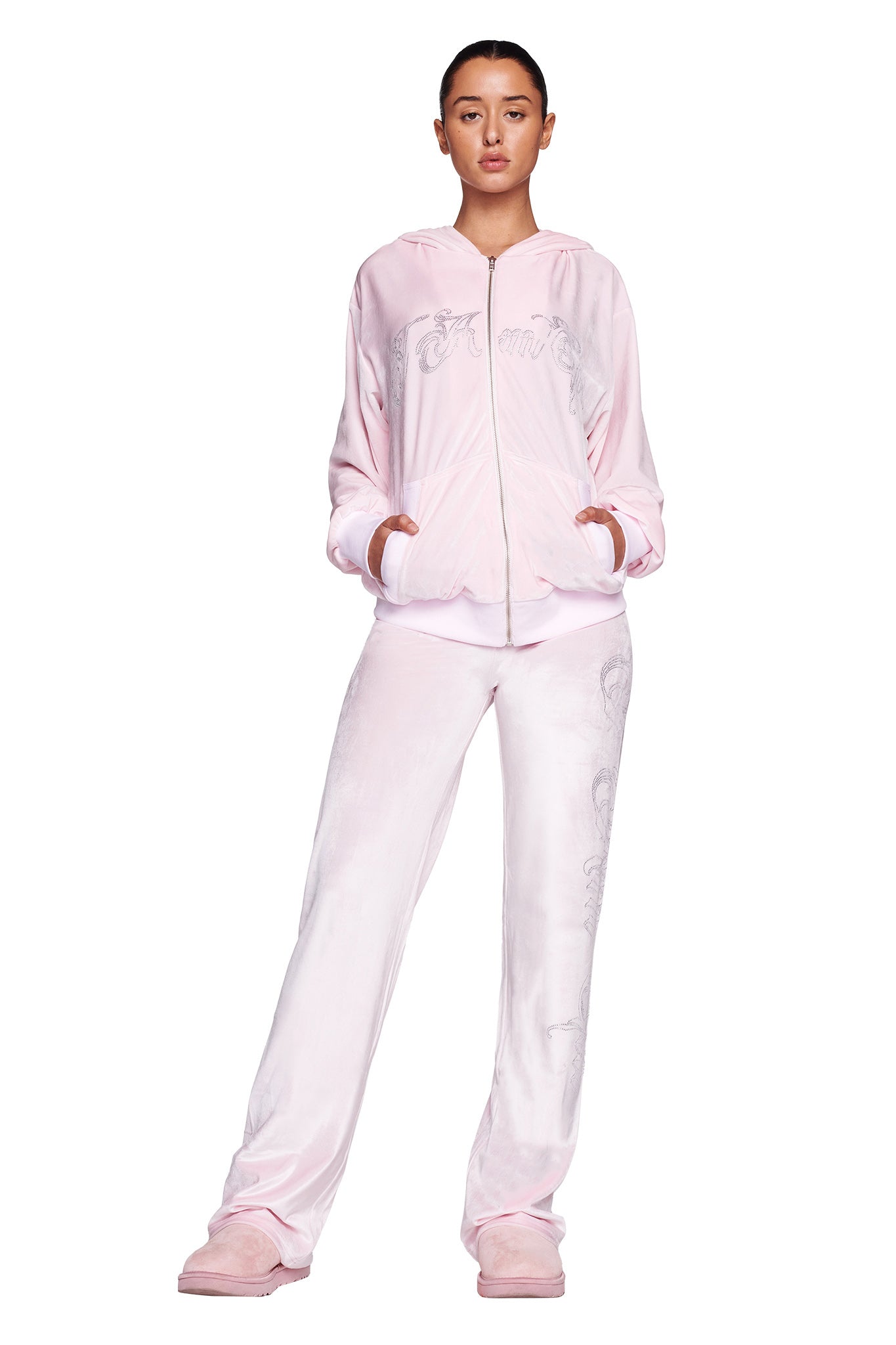 A person stands facing forward, dressed in a light pink hooded zip-up jacket adorned with a custom I.AM.GIA logo and matching DARIA PANT - PINK : ICY PINK from I.AM.GIA. Their hands rest in the jacket pockets, and they wear pink slippers with their hair pulled back. The outfit showcases subtle design details.