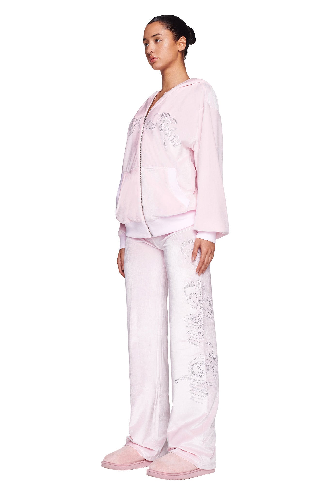 A person stands against a white background wearing the I.AM.GIA DARIA HOODIE in ICY PINK paired with matching wide-leg pants. They are also sporting matching pink slippers, and their hair is pulled back in a neat bun.