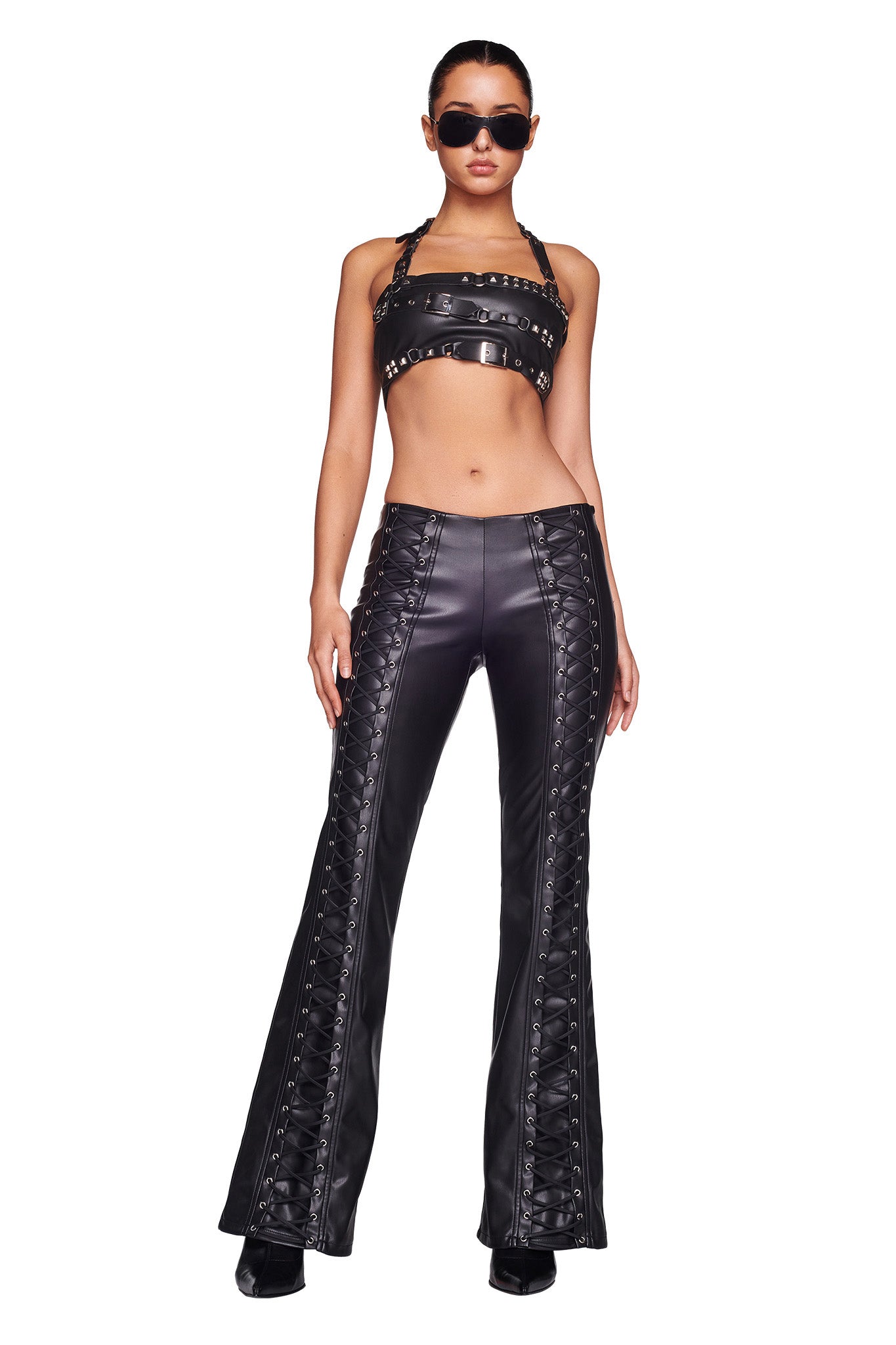 A person wearing the SONORA PANT - BLACK from I.AM.GIA, part of a black leather outfit with a crop top and bell-bottom pants featuring lace-up detailing. They are also wearing dark sunglasses and black shoes, standing against a plain white background.