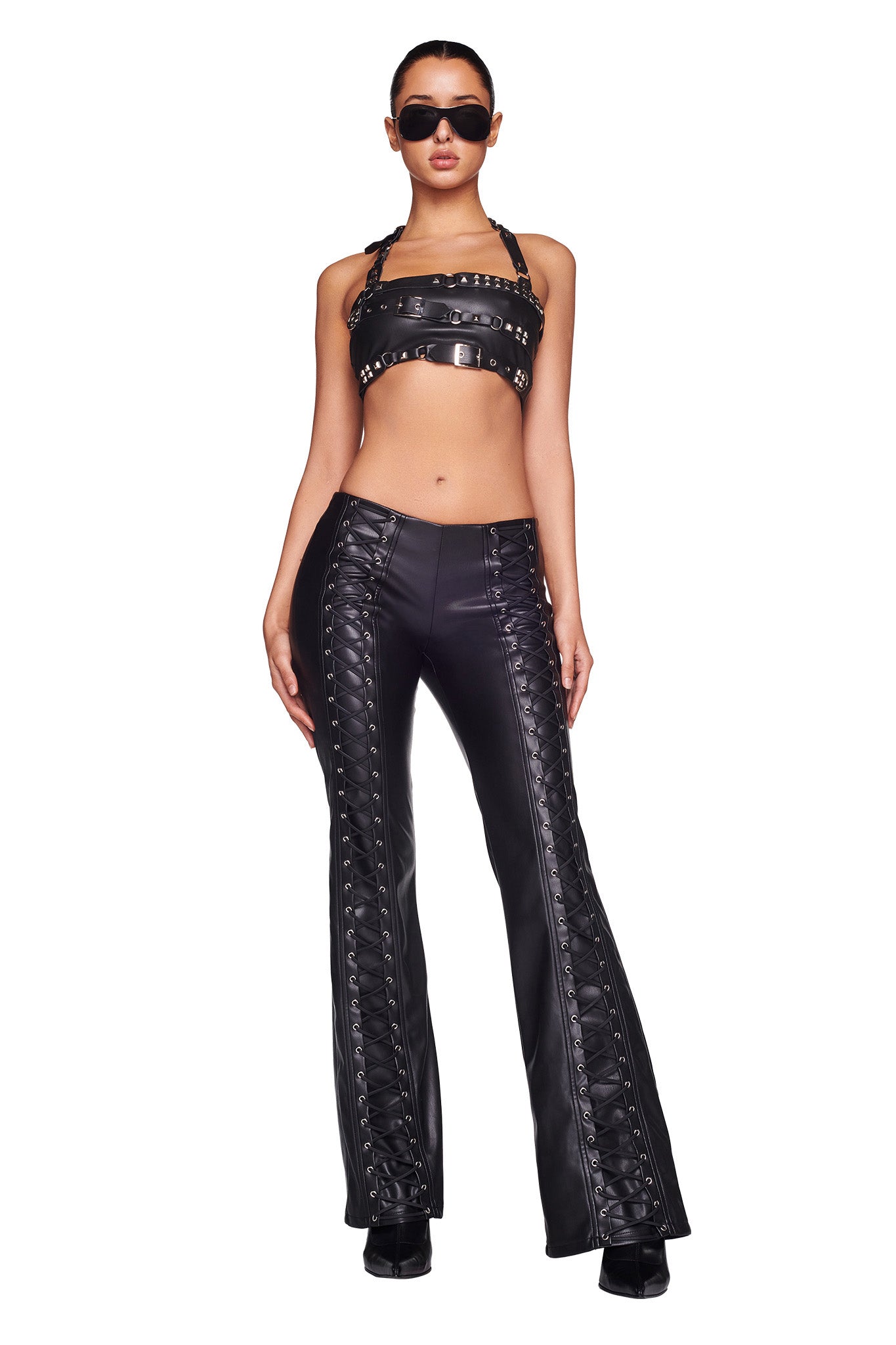 A person wearing a black leather outfit with stud embellishments stands confidently. They are dressed in an I.AM.GIA crop top with straps and the SONORA PANT - BLACK, which features high-waisted flared legs and lace-up details along the sides. They accessorize with black sunglasses and are posing against a white background.