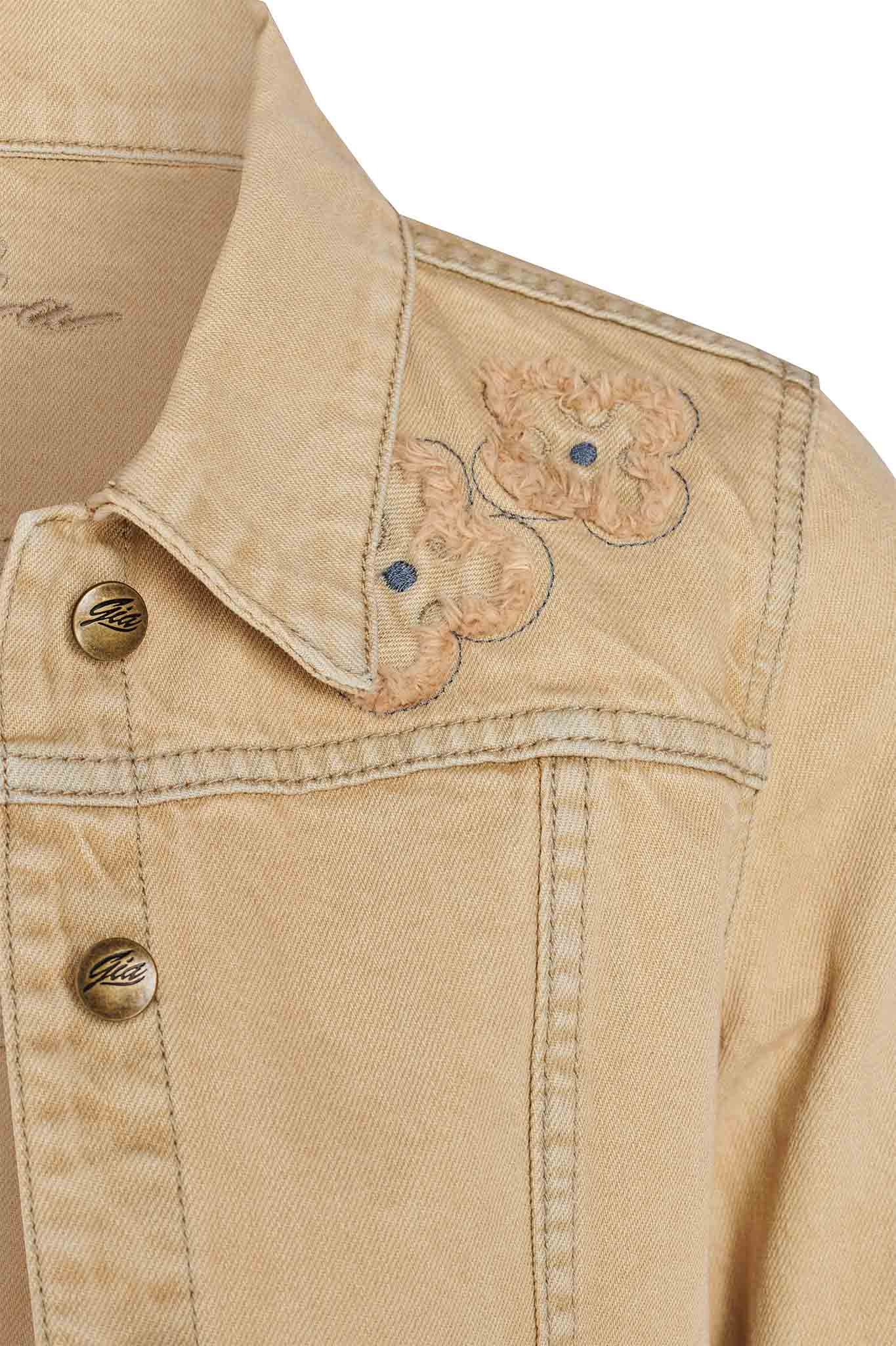 Close-up of the FAUNA JACKET - BEIGE : TAN DENIM by I.AM.GIA showcasing detailed embroidery on the shoulder. The intricate floral designs with blue centers contrast beautifully against the fabric. This jacket features shiny metallic buttons and prominent stitching along the collar and seams, epitomizing chic craftsmanship.