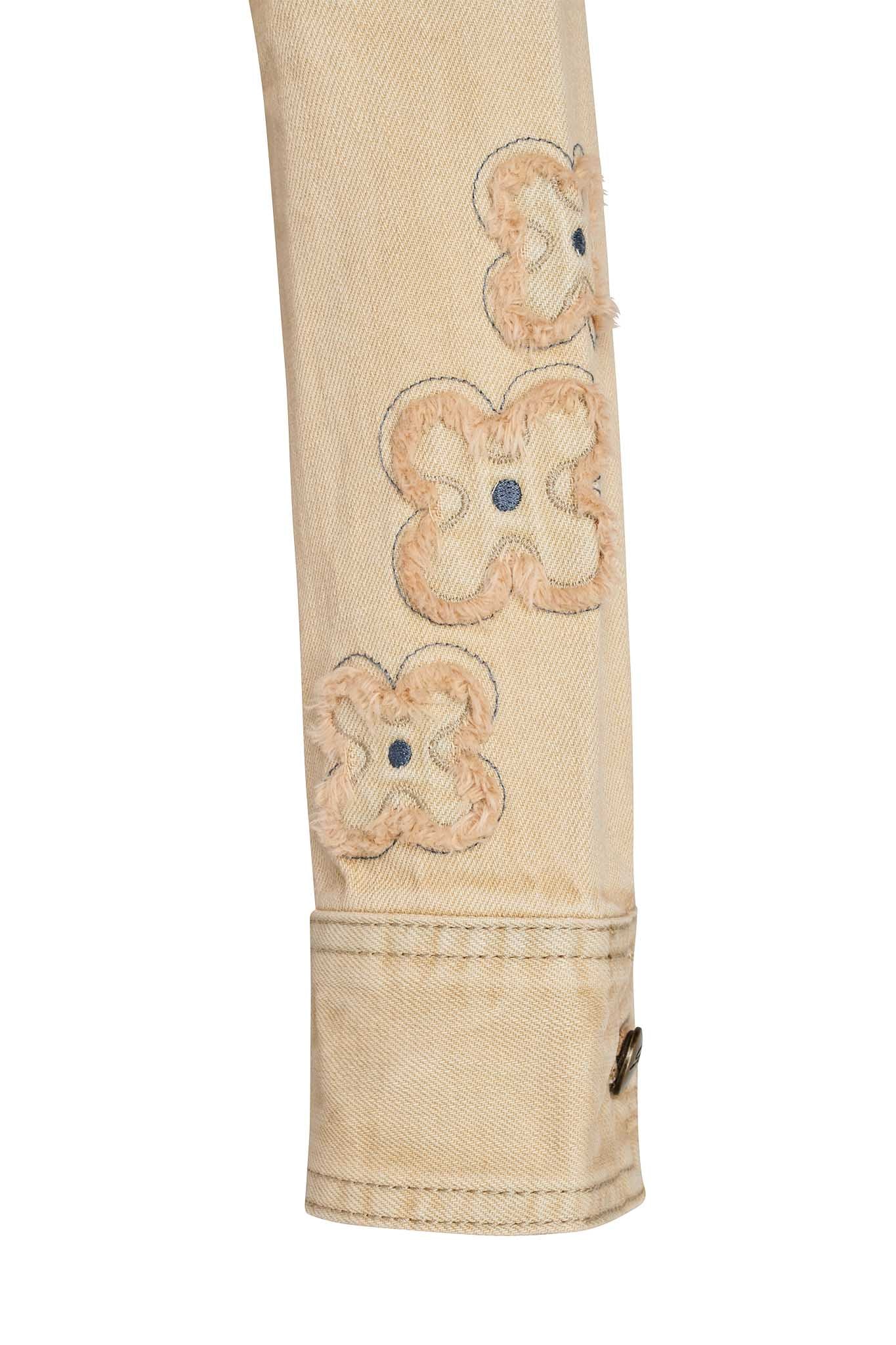 Close-up view of the FAUNA JACKET - BEIGE : TAN DENIM by I.AM.GIA, highlighting the sleeve which features three embroidered, flower-shaped patches in light brown thread with dark center accents. The sleeve is partially turned up, revealing a visible button on the cuff.