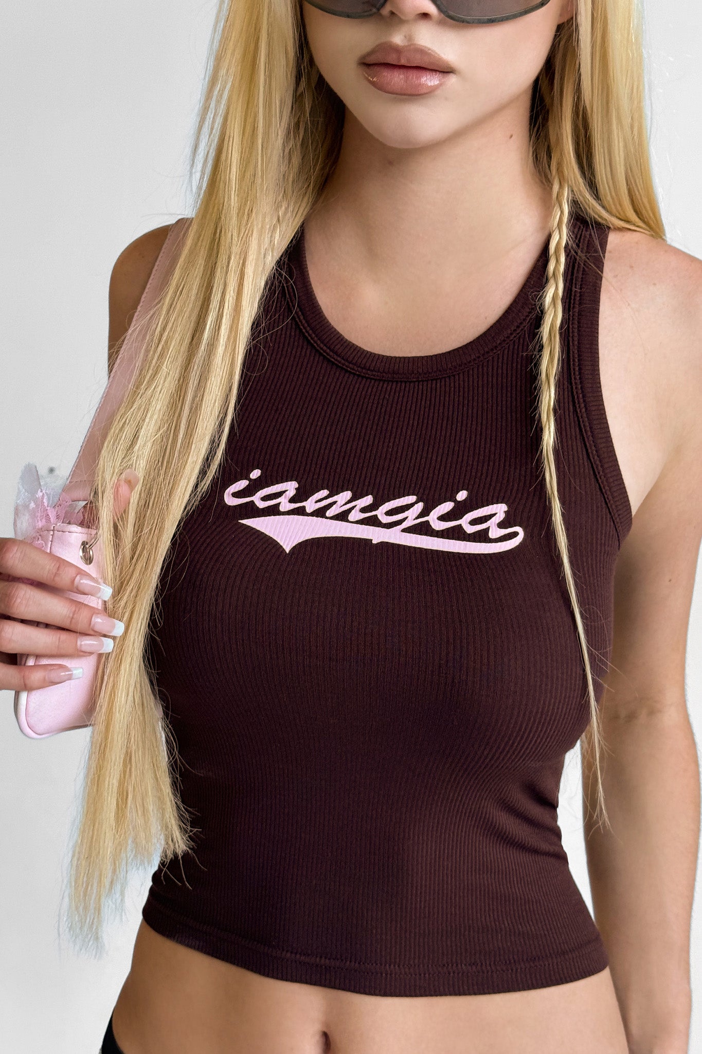 A woman with long blonde hair and sunglasses wears the I.AM.GIA PENELOPE TANK in brown featuring pink "iamgia" script. She carries a pink shoulder bag, and the photo is cropped below her nose.