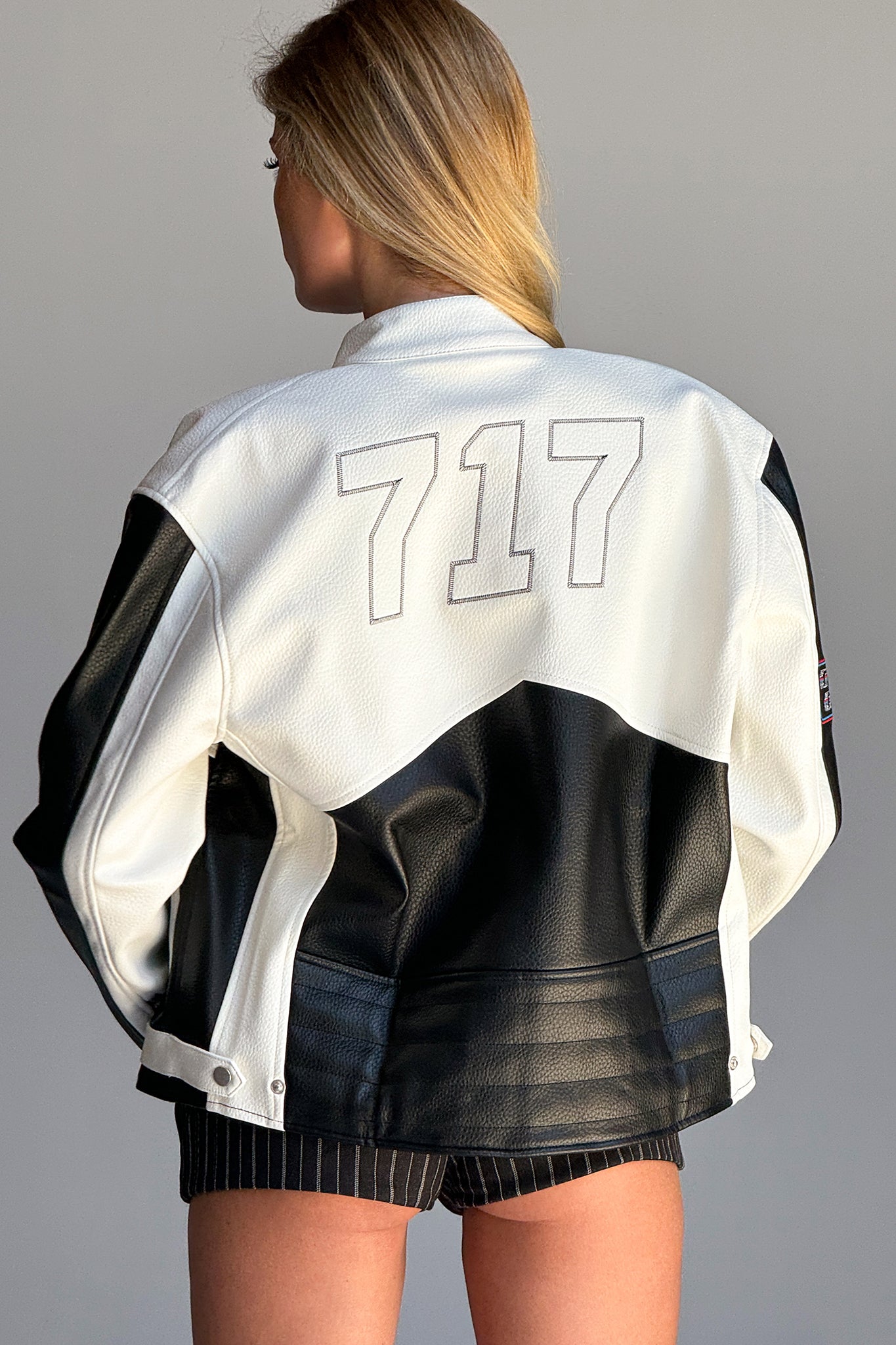 A person with long hair is seen from the back, wearing an oversized I.AM.GIA ROXY JACKET - WHITE with the number "717" prominently displayed on the back. The jacket features a mix of textures, including faux leather, and the person is also wearing dark shorts. The background is plain and light-colored.