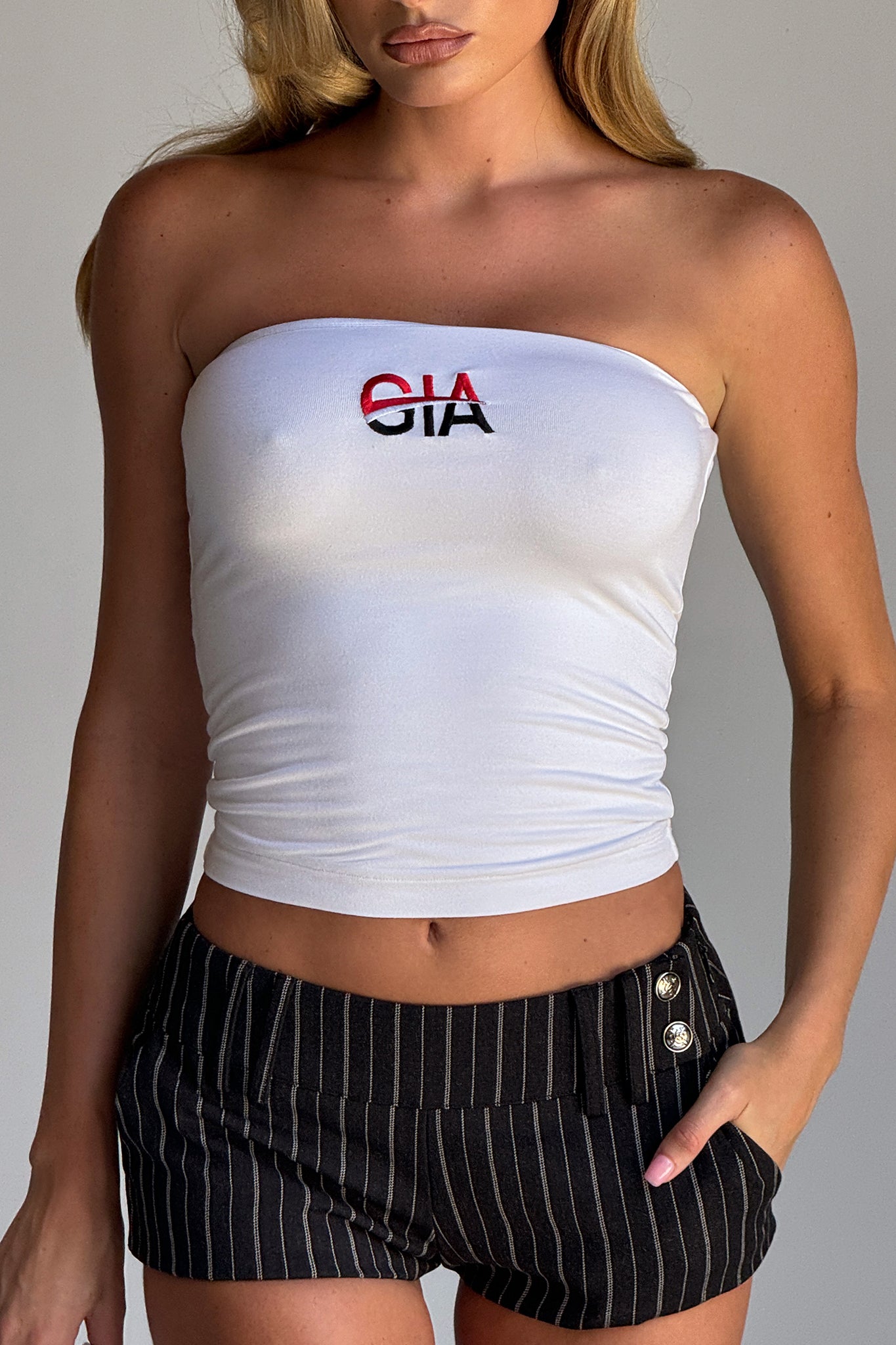 A woman with long blonde hair wears a white strapless tube top emblazoned with "CIA." She pairs it with the SADIE SHORT in black from I.AM.GIA, high-waisted pinstriped suiting shorts featuring button details on the side. The tailored silhouette stands out against a plain, neutral background.