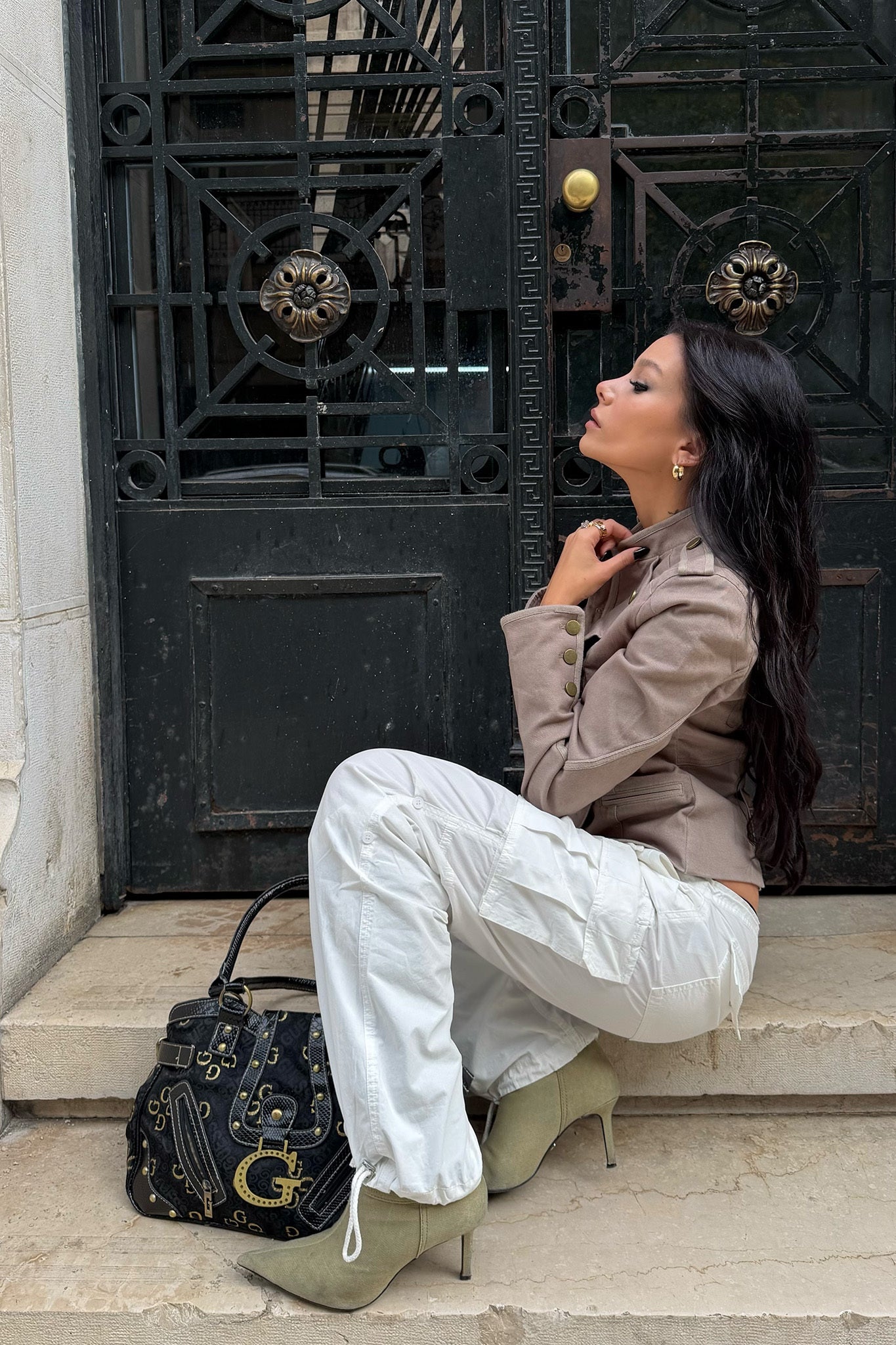 A woman with long dark hair sits on stone steps in front of a black ornate door, wearing I.AM.GIA's IVANA CARGO PANT in white, a taupe jacket, light green heels, and has a black designer handbag beside her.