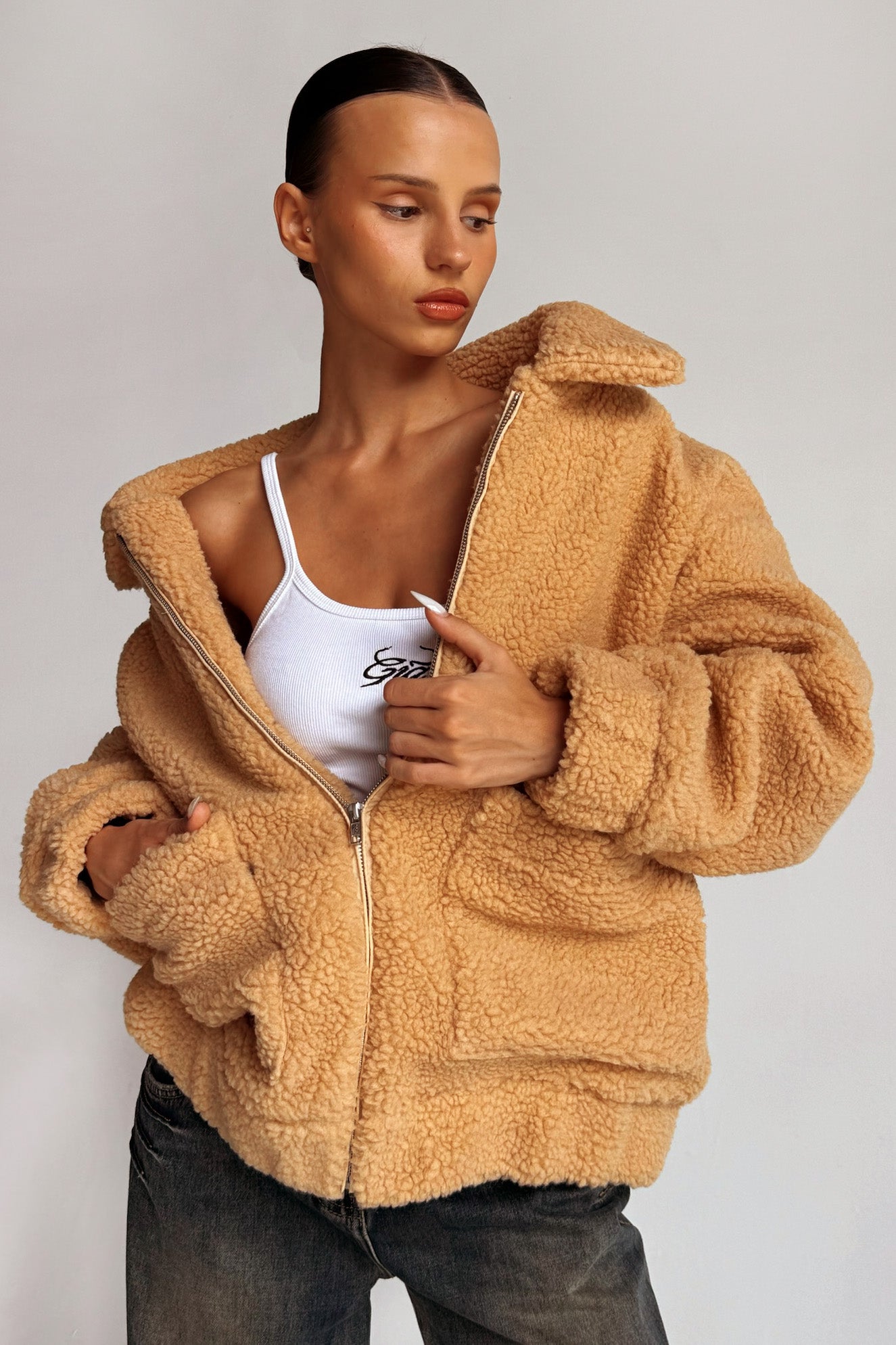 A person with slicked-back hair models the I.AM.GIA PIXIE COAT in caramel, partially unzipped to reveal a white tank top underneath. They are posed with one hand holding the coat and eyes looking away from the camera. Their expression is calm and composed.
