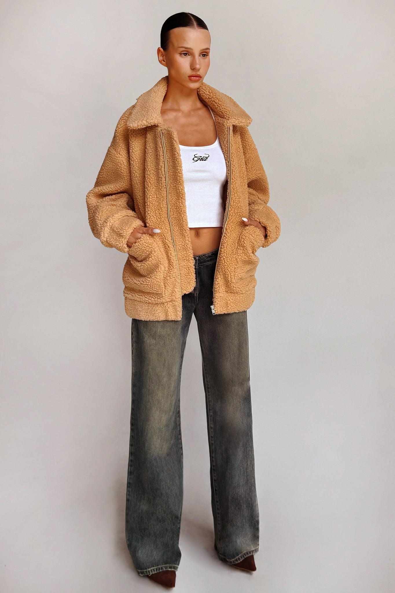 A person stands on a plain background, wearing the I.AM.GIA PIXIE COAT - CARAMEL over a white cropped top and high-waisted, wide-leg gray jeans. Their hands rest in the coat pockets, and they wear pointed brown shoes. They have a neat hairstyle and a composed expression.