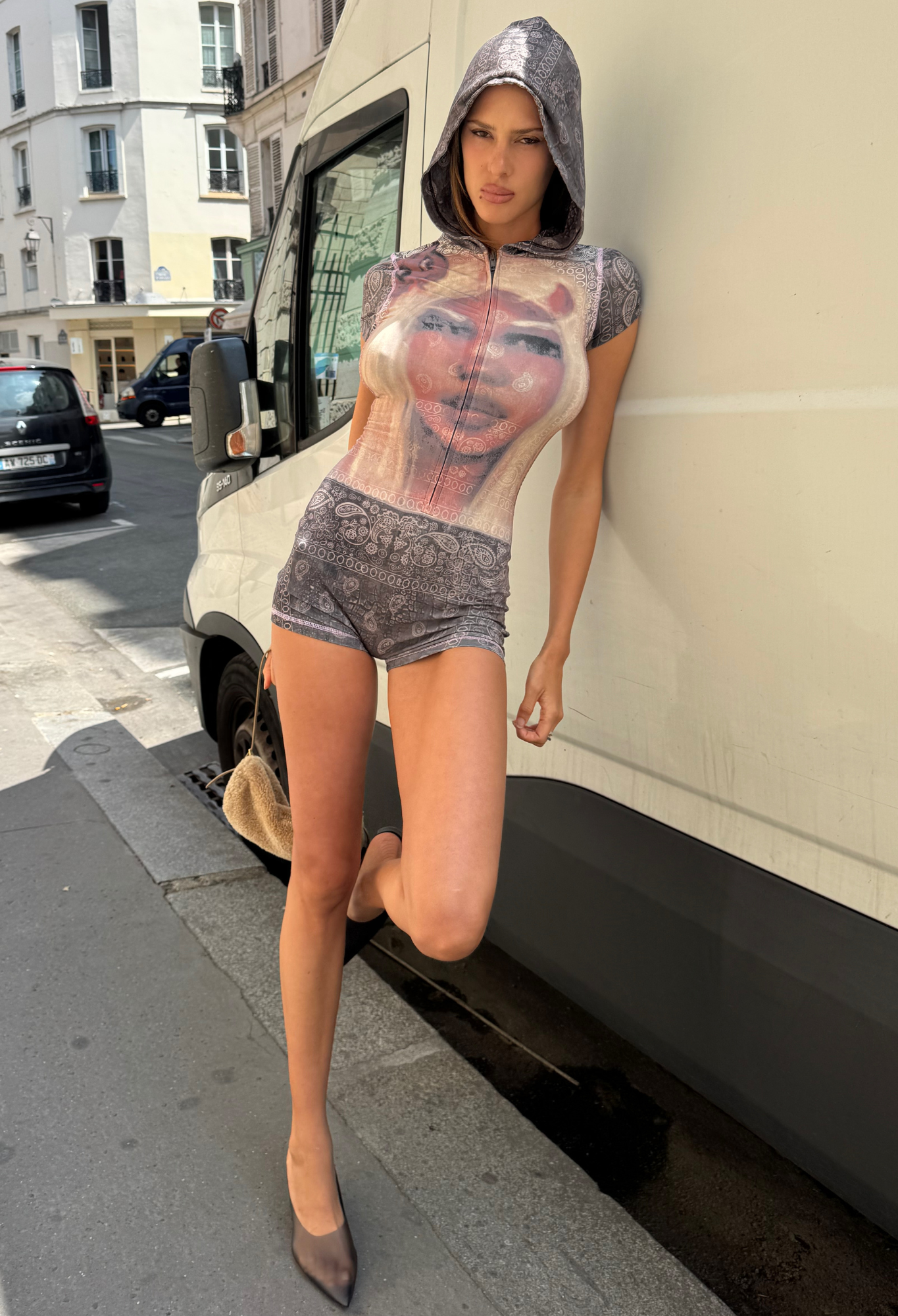 Wearing the I.AM.GIA T-REX BLARE SHORT SLEEVE ROMPER in Charcoal Bandana, a person stands against a white van on a city street, one leg bent, styled with pointed heels and a small bag—a bold and edgy festival look.