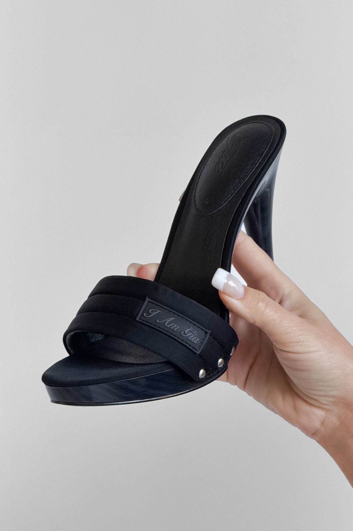 A hand with manicured nails holds the NICKY HEEL - BLACK NEOPRENE by I.AM.GIA, a black slip-on mule sandal with a thick front strap and stiletto heel, set against a plain light background.