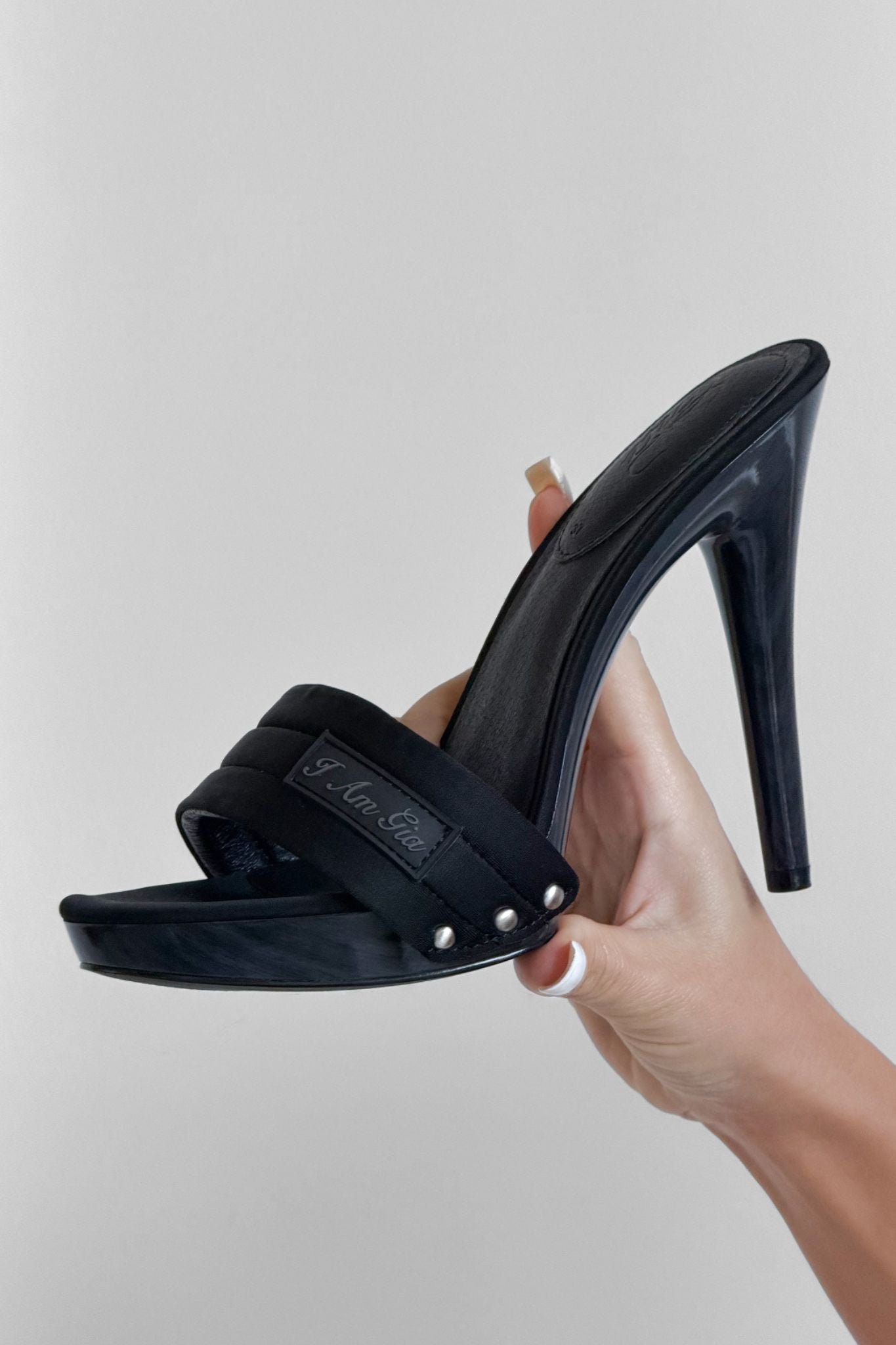 A manicured hand holds the I.AM.GIA NICKY HEEL in black neoprene, showcasing its platform sole, open toe, padded vamp strap, and bold rivet accents against a plain light background.