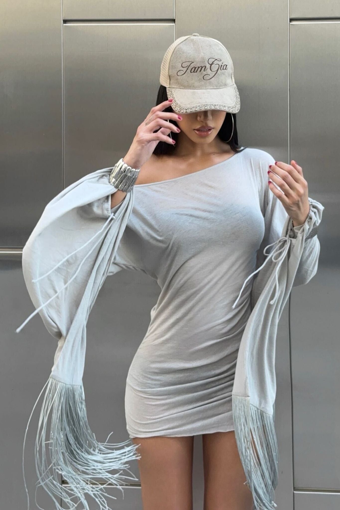 A woman in the I.AM.GIA LUMA MINI DRESS - GREY with fringe sleeves and a beige cap chats on her cell phone, one hand on her hip, against a metallic background.