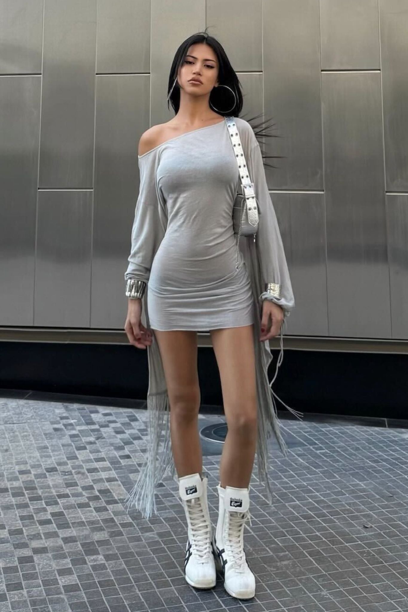 A woman poses outdoors in the I.AM.GIA LUMA MINI DRESS - GREY, styled with white lace-up boots, large hoop earrings, and a white crossbody bag, set against a metallic wall and tiled ground.