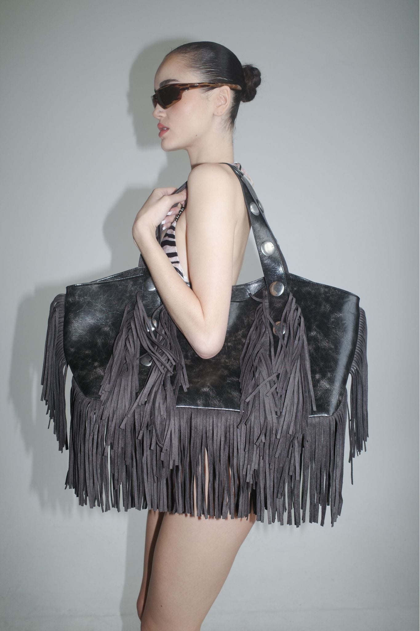 A woman in sunglasses and a striped swimsuit poses sideways, holding the I.AM.GIA CHROMA FRINGE BAG - BLACK, an oversized faux leather tote with distressed black fringe and large handles, against a plain light background.