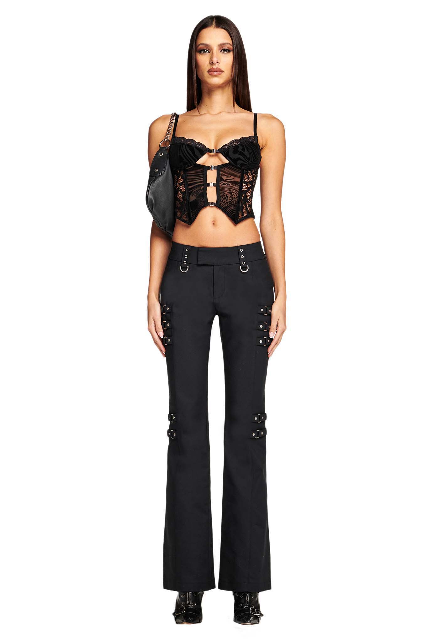 A woman stands against a white background. She wears black, flared pants with silver buckles and an I.AM.GIA ELENA TOP - BLACK BURNOUT lace-up crop top. She carries a small black purse over her shoulder and wears black, high-heeled boots. Her long, straight hair is parted down the middle.