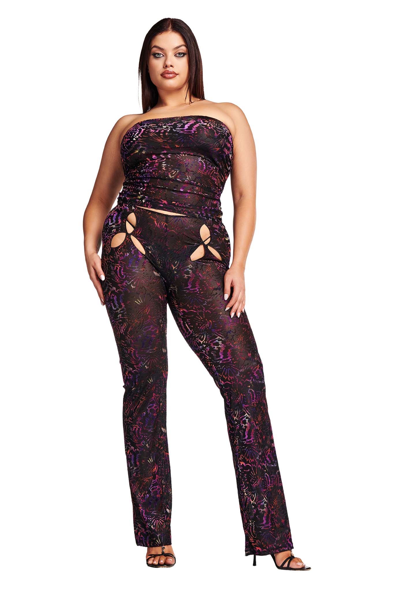 A woman stands confidently in I.AM.GIA's AMINA PANTS - ANIMAL BURN OUT, a colorful jumpsuit with abstract patterns of purple, pink, and black. The outfit features stylish cut-out details near the hips. She wears black high-heeled sandals and has long, dark hair cascading down her back.