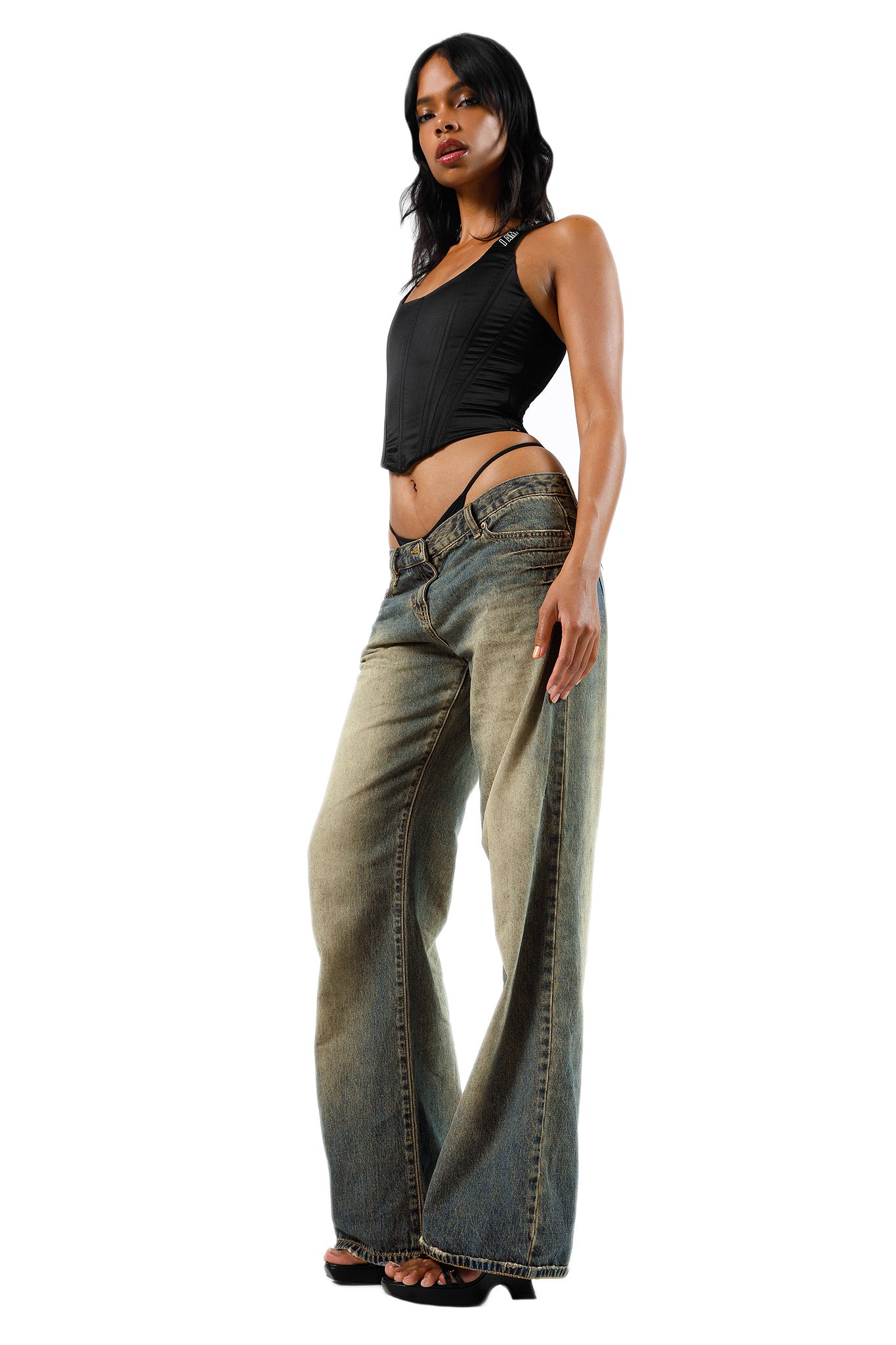 A person posing in a black, sleeveless I.AM.GIA SIOBHAN CORSET with a front zipper and distressed, loose-fitting jeans. They have long, dark wavy hair and are standing with one hand on their hip, exhibiting a confident stance against a plain white background.