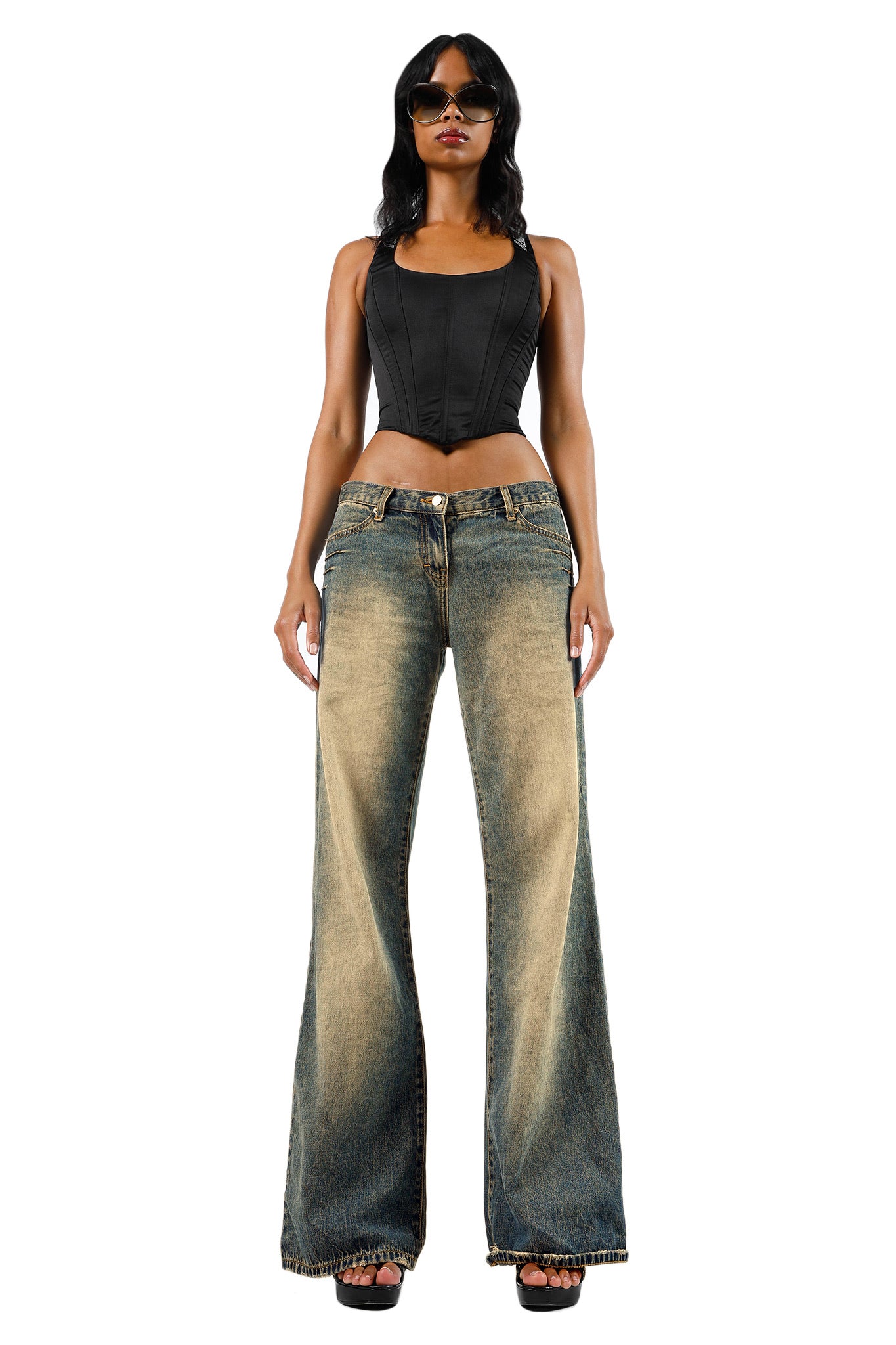 A woman wearing I.AM.GIA sunglasses, the SIOBHAN CORSET - BLACK by I.AM.GIA, and wide-legged faded blue jeans stands confidently against a plain white background. She completes her look with black high-heeled shoes.