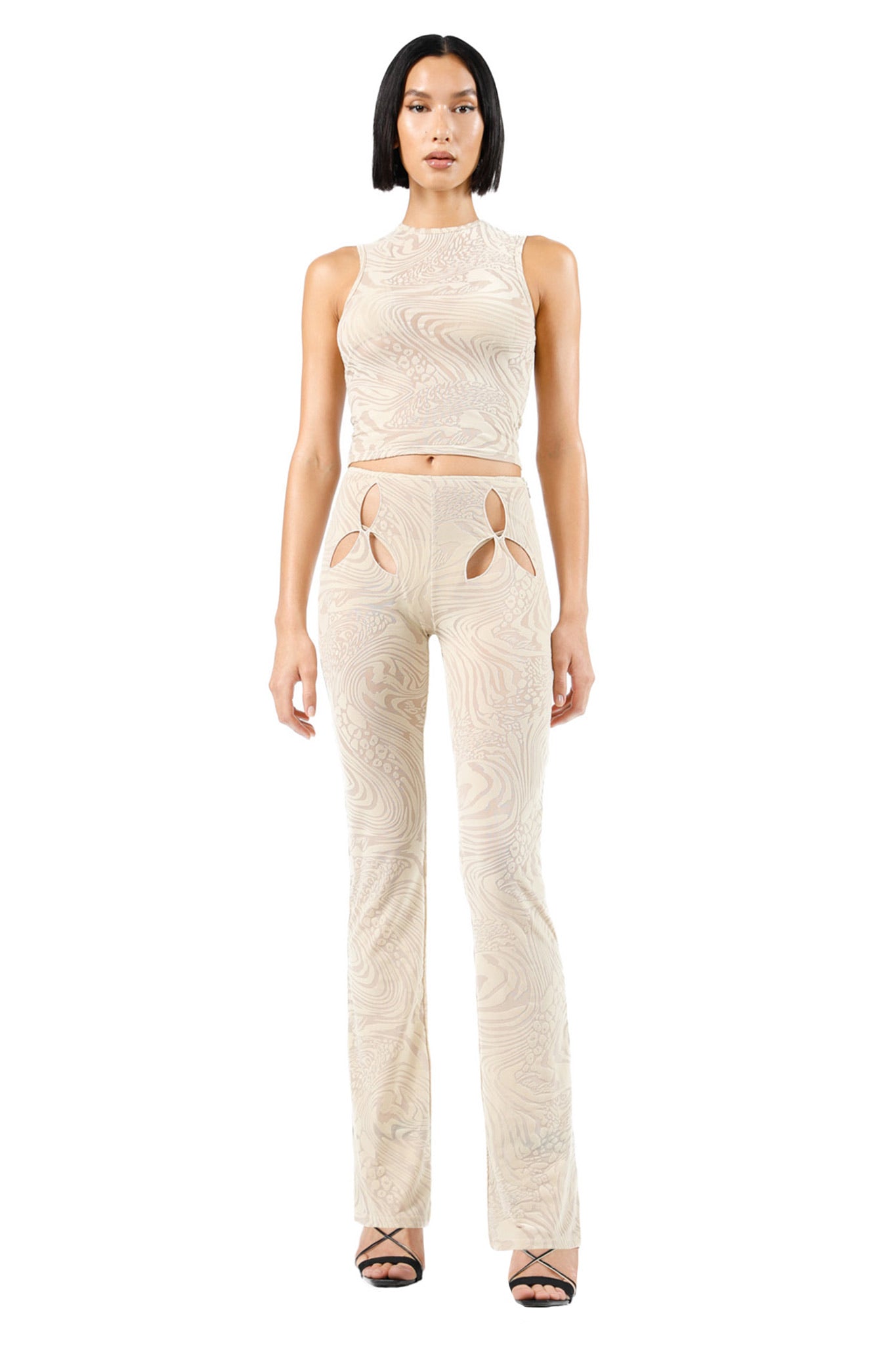 A woman stands facing the camera wearing the AMINA TOP - TAN BURNOUT from I.AM.GIA, a cream-colored, sleeveless crop top paired with matching high-waisted pants featuring artistic patterns. The pants have cut-outs around the upper thigh area, and she completes her look with strappy high-heeled sandals.