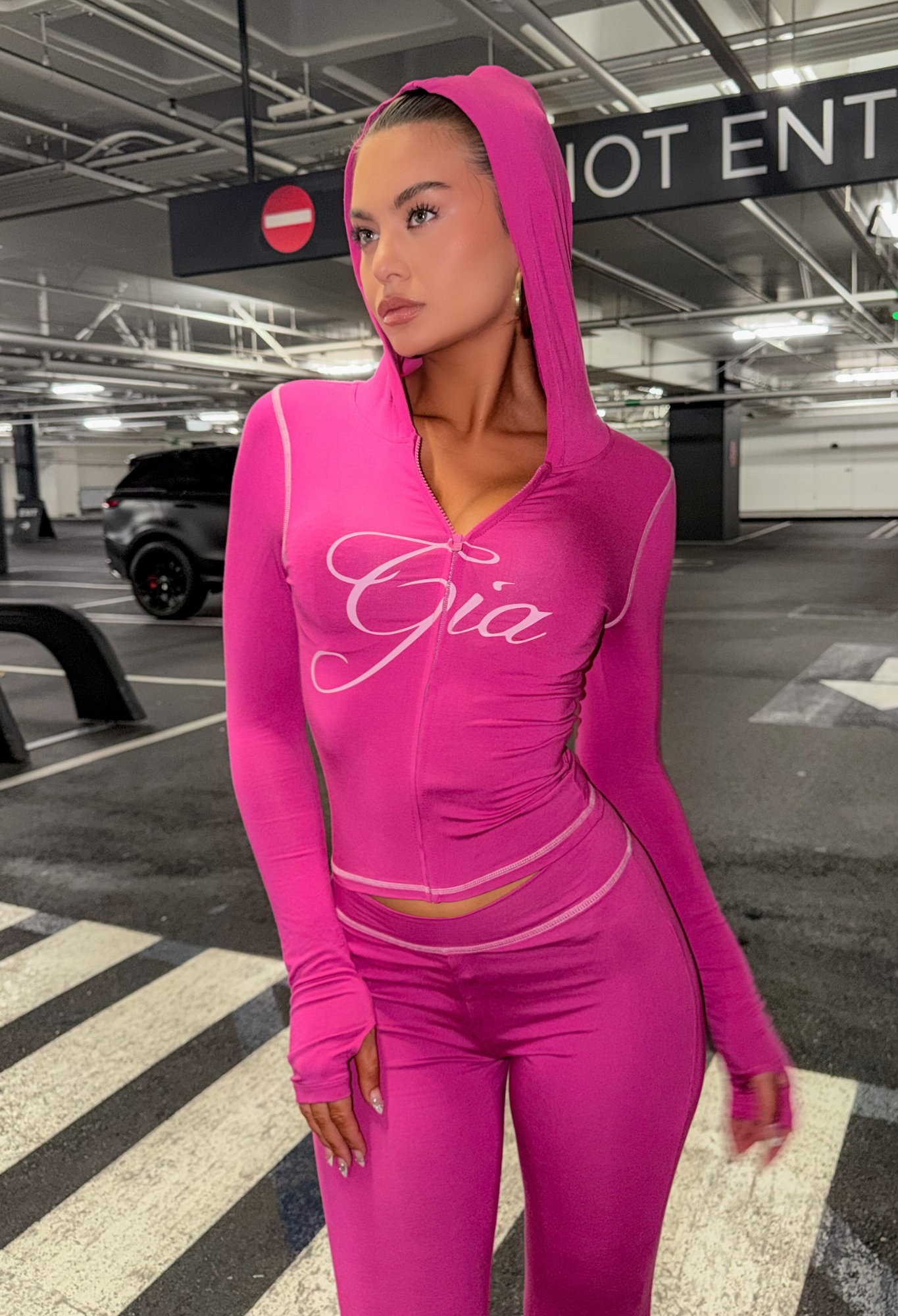 A woman wearing the I.AM.GIA BLARE HOODIE in Raspberry stands in a parking garage near a pedestrian crossing, gazing to the side. A "Do Not Enter" sign and several parked cars appear in the background.