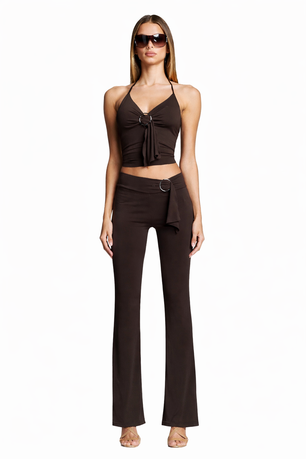 A woman faces forward, barefoot, wearing sunglasses and the I.AM.GIA SIOFRA PANT in brown with a side ring detail, complemented by a sleeveless brown halter top with a front tie. She poses against a plain white background.