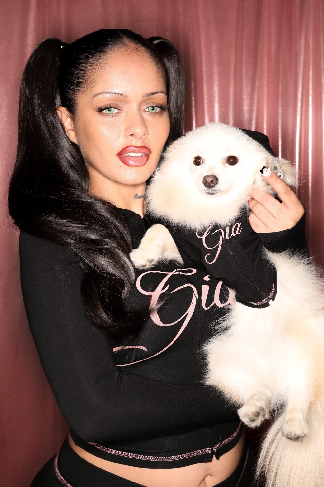A woman with long dark hair in pigtails poses with her fluffy white dog in front of a pink curtain. Both wear matching I.AM.GIA BLARE BEST FRIEND DOG HOODIE - BLACK featuring pink writing. She has light eyes and glossy lips.