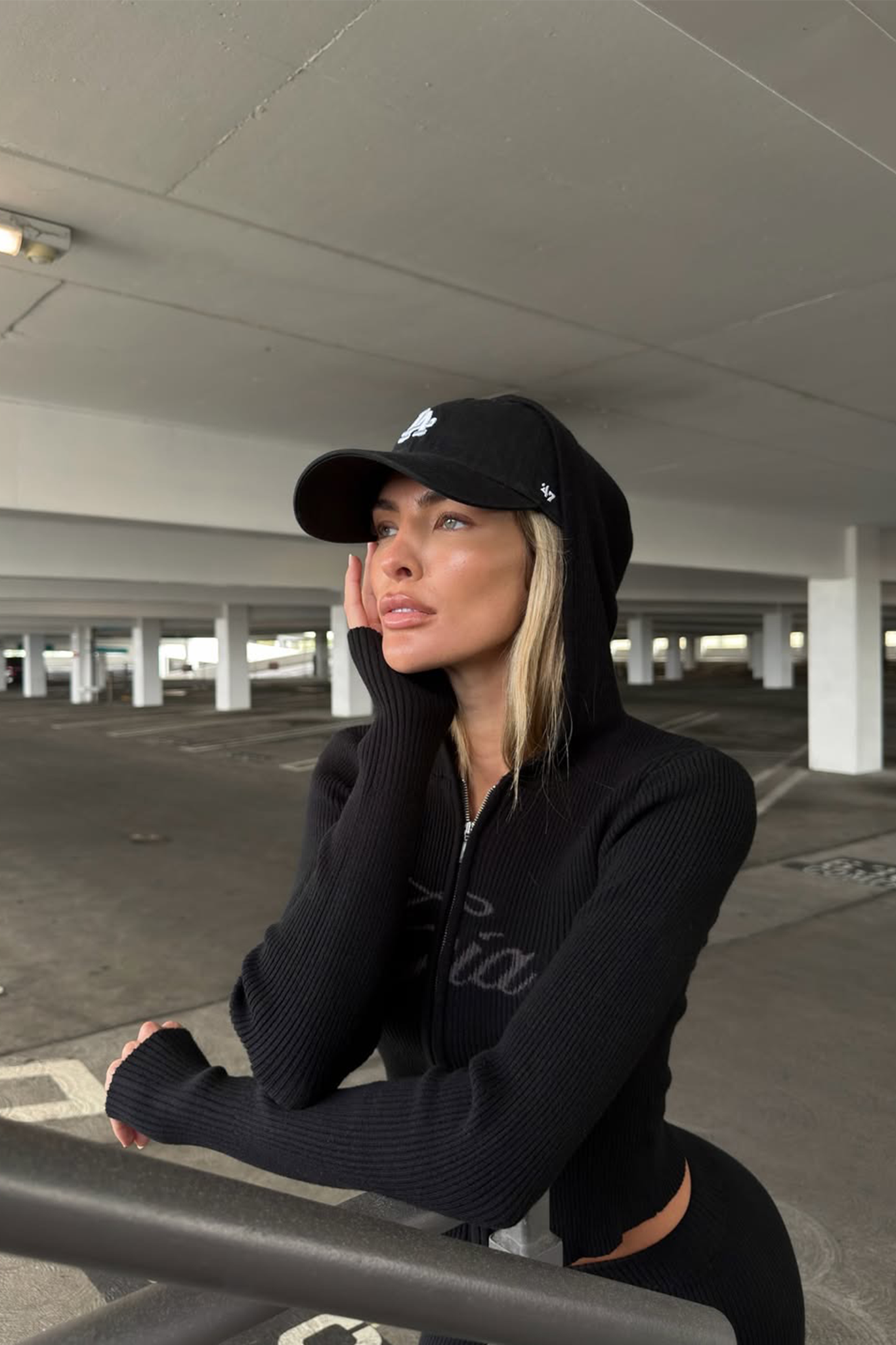 A woman in the I.AM.GIA BLARE KNIT HOODIE - BLACK and a black cap leans on a metal railing in a parking garage, gazing thoughtfully into the distance with her hand on her face, embodying I.AM.GIA’s iconic style.