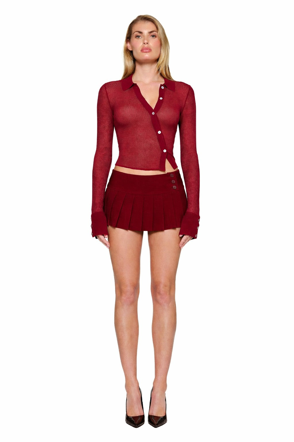 A woman with straight blonde hair wears the I.AM.GIA DAVINA TOP in wine red, a sheer knit, long-sleeved button-up, paired with a matching short pleated skirt and black high heels on a white background.