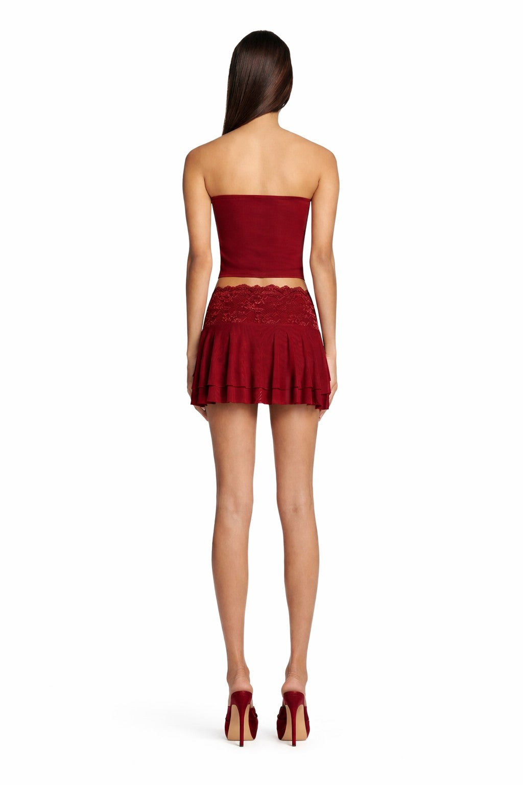 A woman with long, straight brown hair stands facing away against a plain white background, wearing the I.AM.GIA DEMIE SKIRT in deep red and a matching strapless crop top with high-heeled shoes.