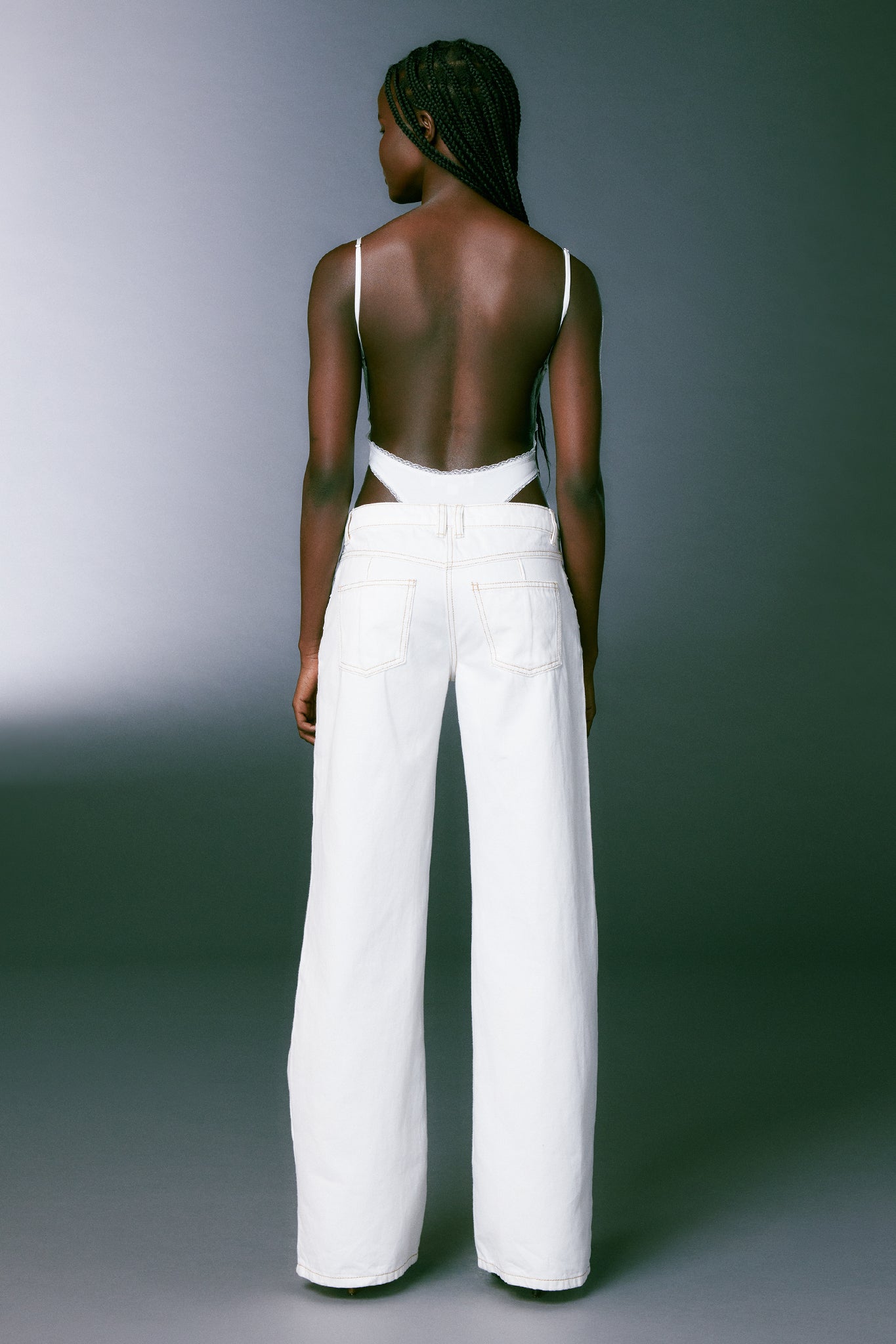 A person stands with their back to the camera, showcasing their I.AM.GIA outfit. They are wearing a backless white top and the DYLAN JEAN - WHITE, which features high-waisted, wide-leg pants. The background is a gradient from dark grey to light grey, creating a simple and minimalist setting.