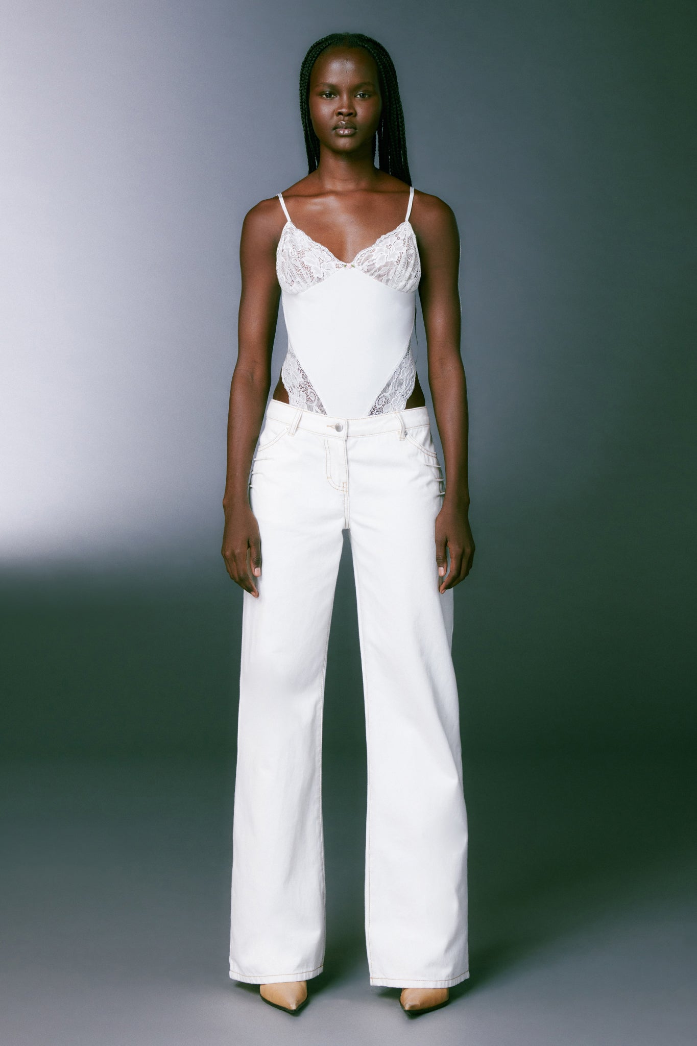 A woman stands against a gray background, dressed in a white camisole with lace details and I.AM.GIA's DYLAN JEAN - WHITE high-waisted wide-leg pants. She is wearing beige pointy shoes from I.AM.GIA. Her hair is styled in long braids, and her expression is calm and focused towards the camera.