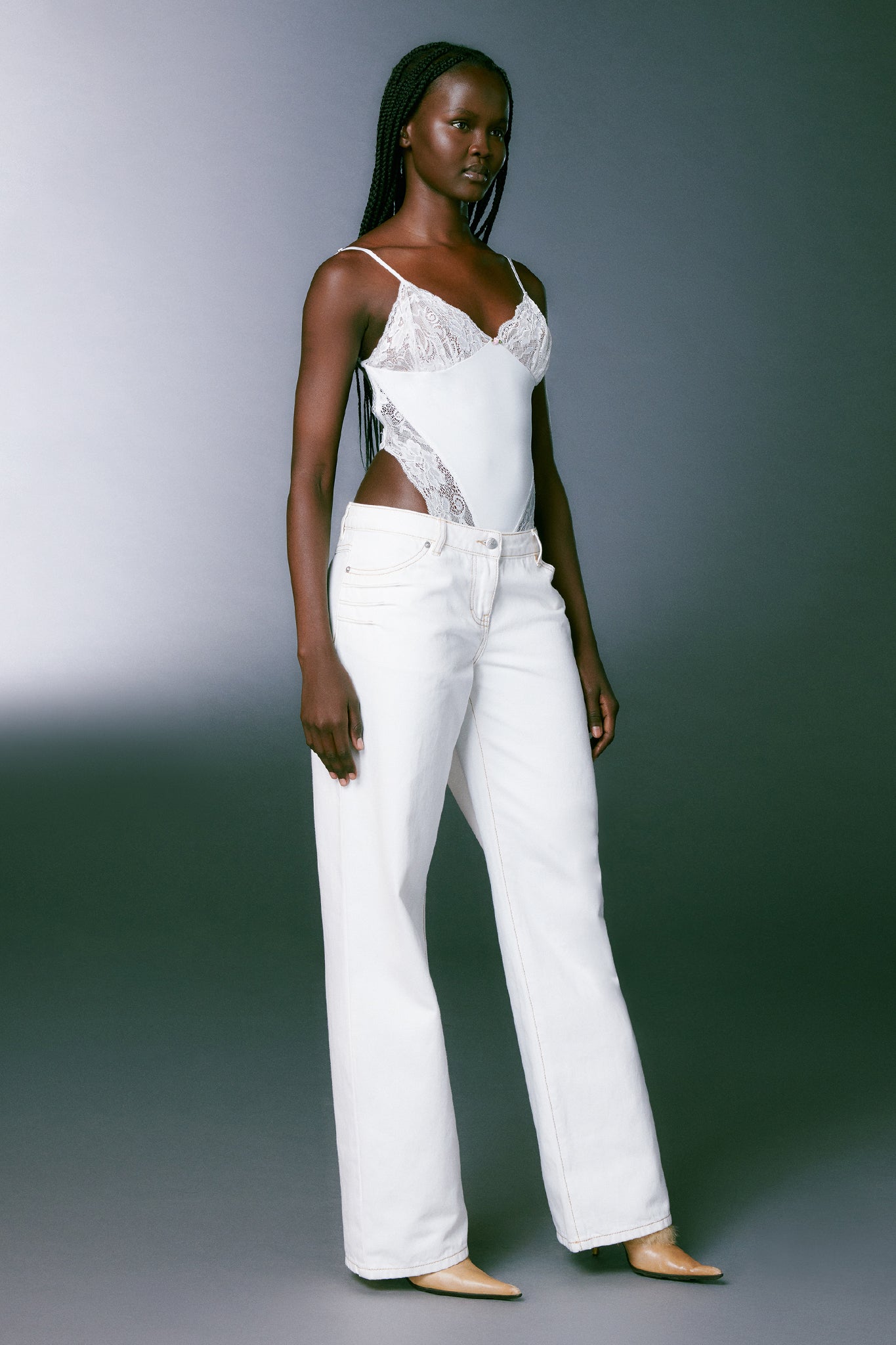 A person with long braided hair wears a white lace bodysuit and the DYLAN JEAN in white by I.AM.GIA. They stand confidently against a grey gradient background, with a neutral expression and hands resting by their sides. The overall look is stylish and minimalist.