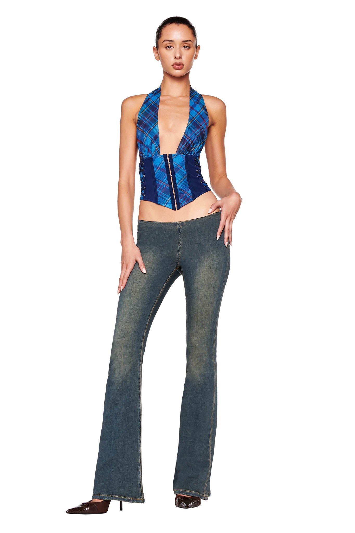 A person with long hair tied back is posing confidently in the CELESTINA CORSET - BLUE : PLAID from I.AM.GIA and high-waisted flared denim jeans. They are wearing dark-colored heeled shoes and standing against a white background.