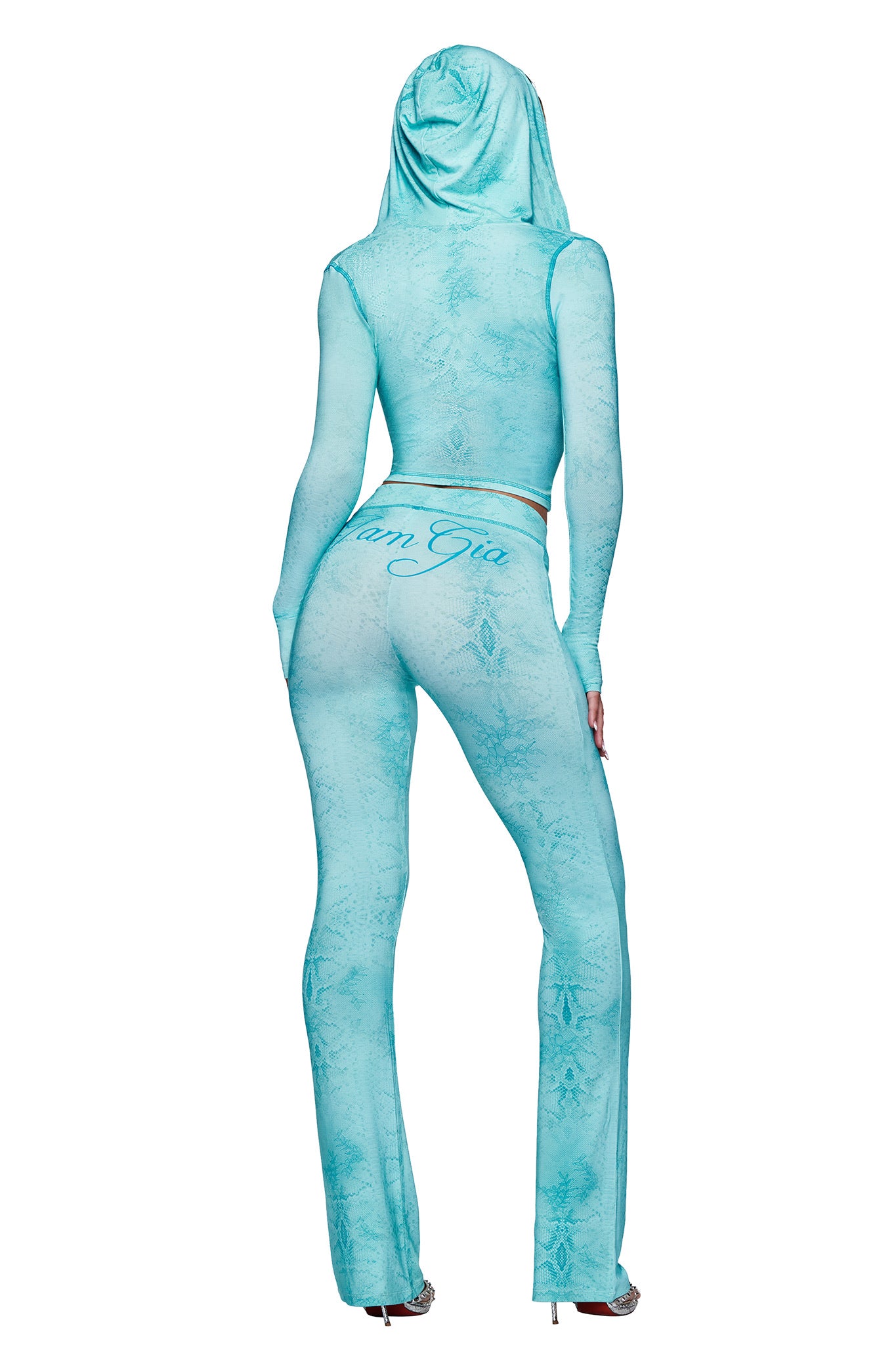 Wearing I.AM.GIA’s BLARE TRACKPANT - TEAL SERPENTINE, a person faces away in a fitted sheer teal hooded outfit with flared legs and long sleeves, styled with heels for a bold statement.