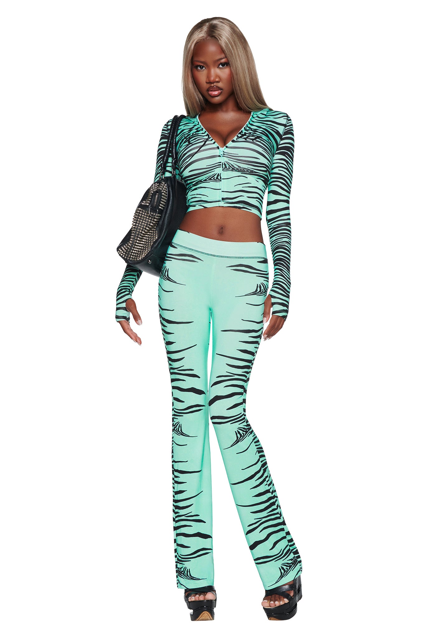 A woman with long straight hair wears the I.AM.GIA BLARE HOODIE in Aqua Fine Line Zebra, paired with matching flared pants, black heels, and a black textured handbag. She stands confidently against a white background.