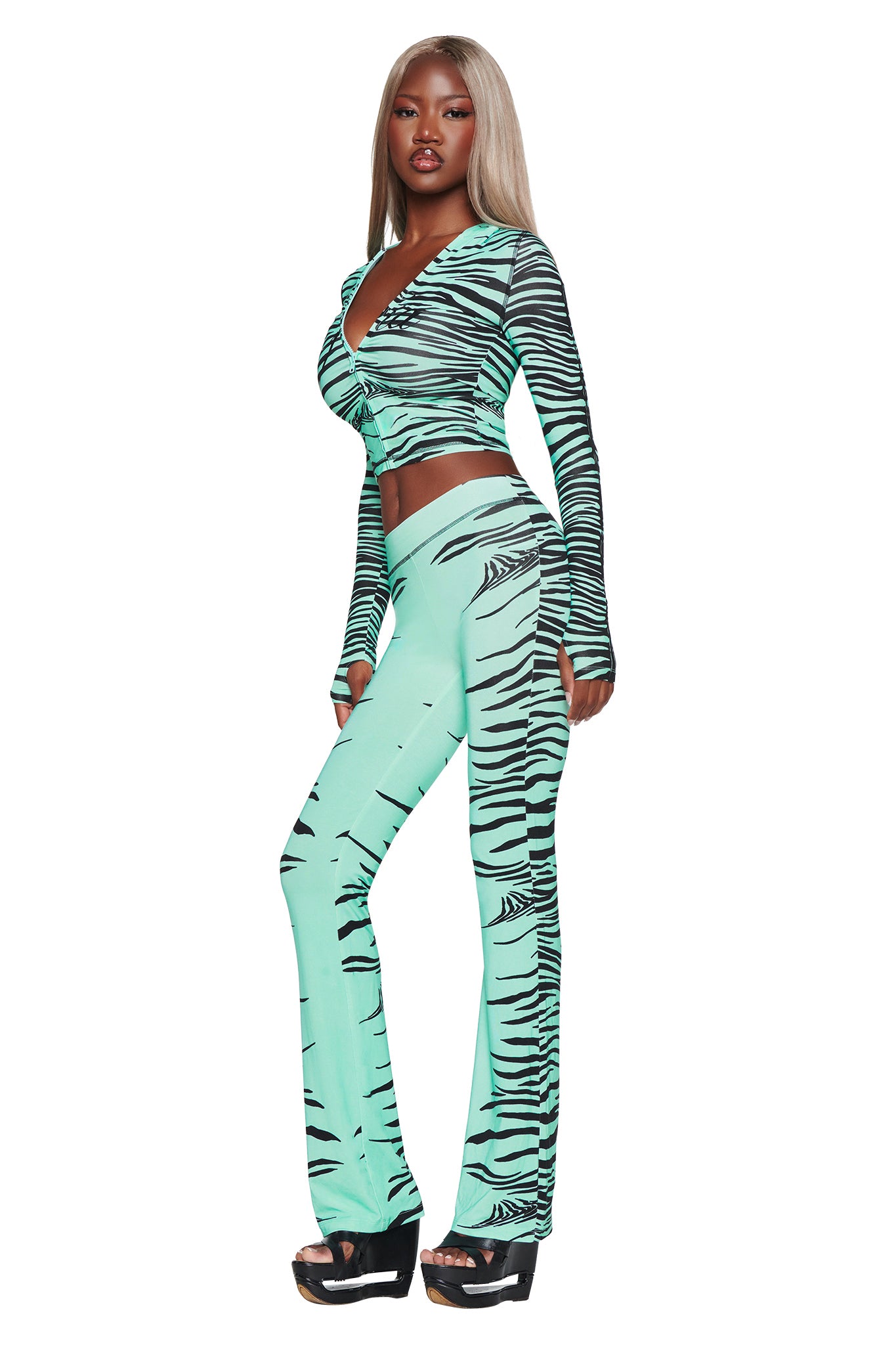 Dressed in the I.AM.GIA BLARE HOODIE - AQUA FINE LINE ZEBRA, a woman stands against a white background. The mint green and black zebra print hoodie, crafted from premium bamboo fabrication, complements her long, straight blonde hair. She completes the look with bell-bottom pants and platform sandals.