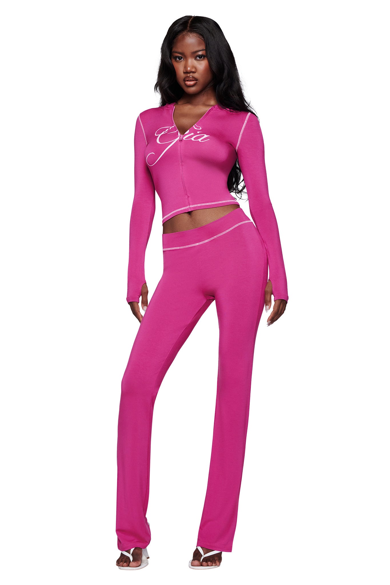 A woman exudes confidence in a vibrant two-piece with a long-sleeve crop top featuring white piping and chest text, reminiscent of the bold I.AM.GIA BLARE HOODIE - RASPBERRY. She pairs it with high-waisted pants and white sandals for a standout look.
