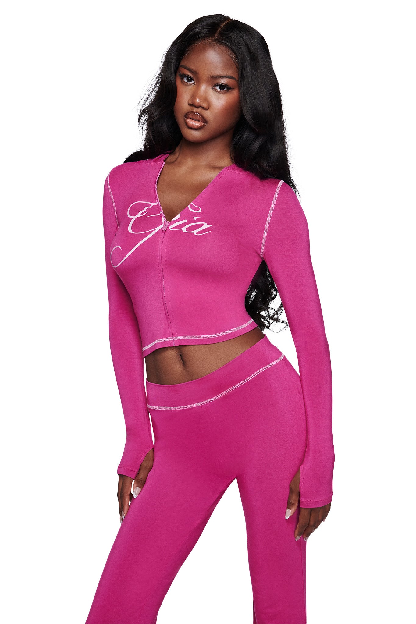 A woman models the I.AM.GIA BLARE HOODIE in raspberry, a bright pink long-sleeve top featuring white stitching and "Gia" script, paired with high-waisted pants for a bold two-piece look.