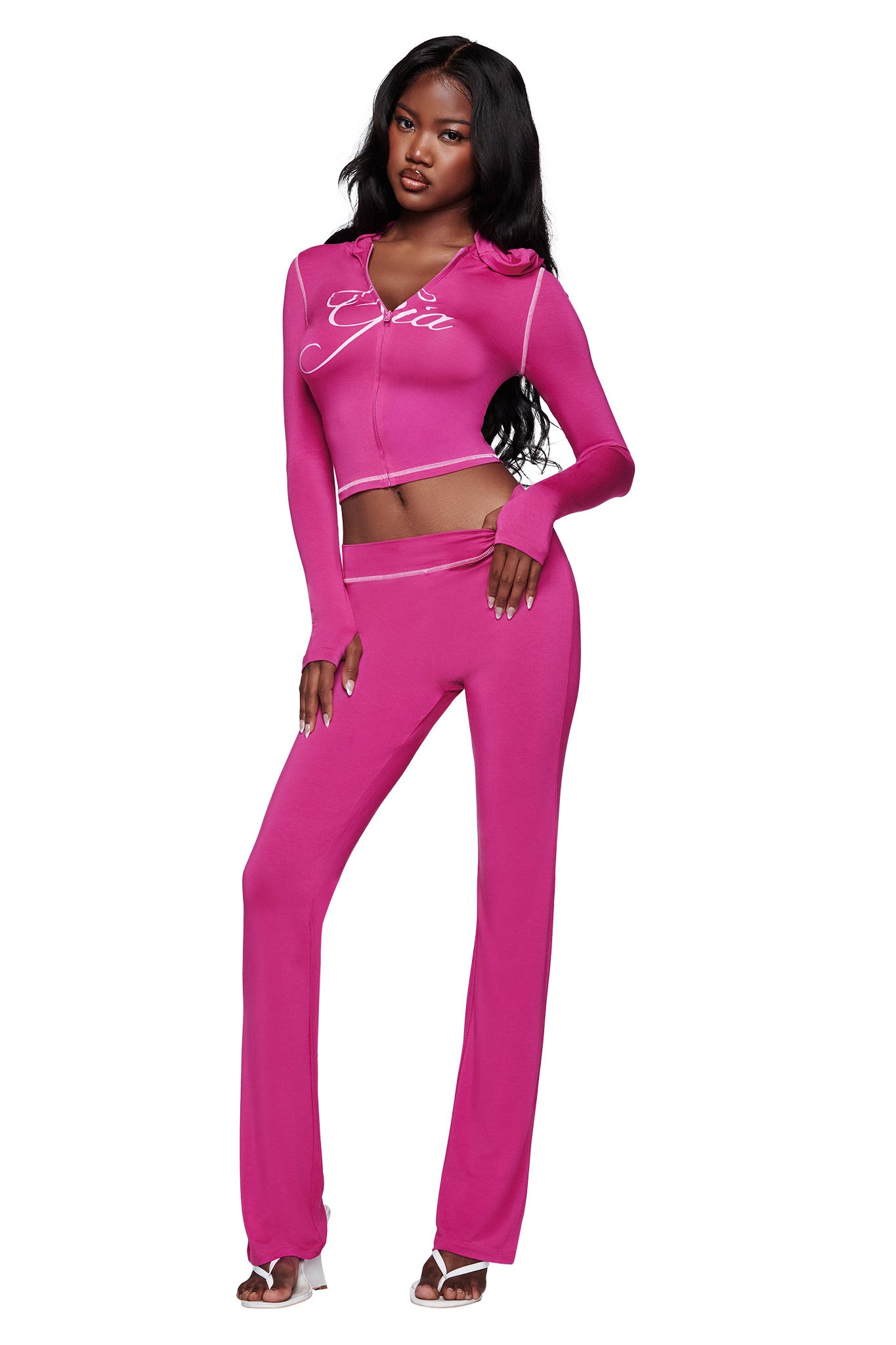 A woman poses confidently in the I.AM.GIA BLARE HOODIE - RASPBERRY, a bright pink two-piece set with long sleeves and flared pants, featuring a hood and cursive front detail. She styles it with white heeled sandals and long hair.