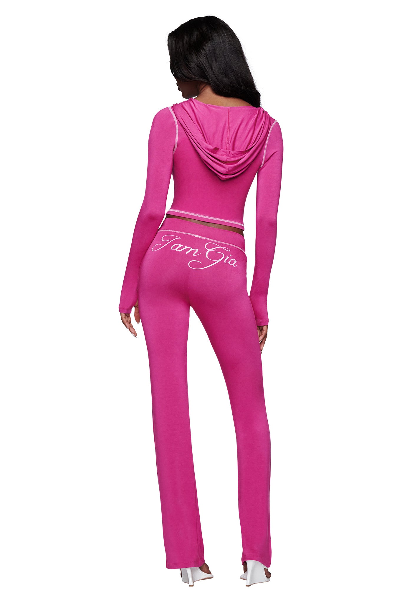 A woman with long dark hair faces away, wearing the I.AM.GIA BLARE HOODIE in raspberry with "IamGia" in white script on the back, paired with matching tracksuit bottoms and white heeled sandals.