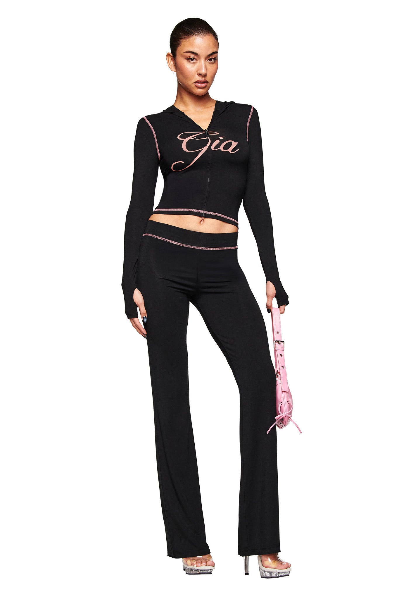 A woman poses in the I.AM.GIA BLARE HOODIE - BLACK with "Gia" on the chest, paired with black flared pants and silver platform heels, holding a pink handbag against a plain white background.