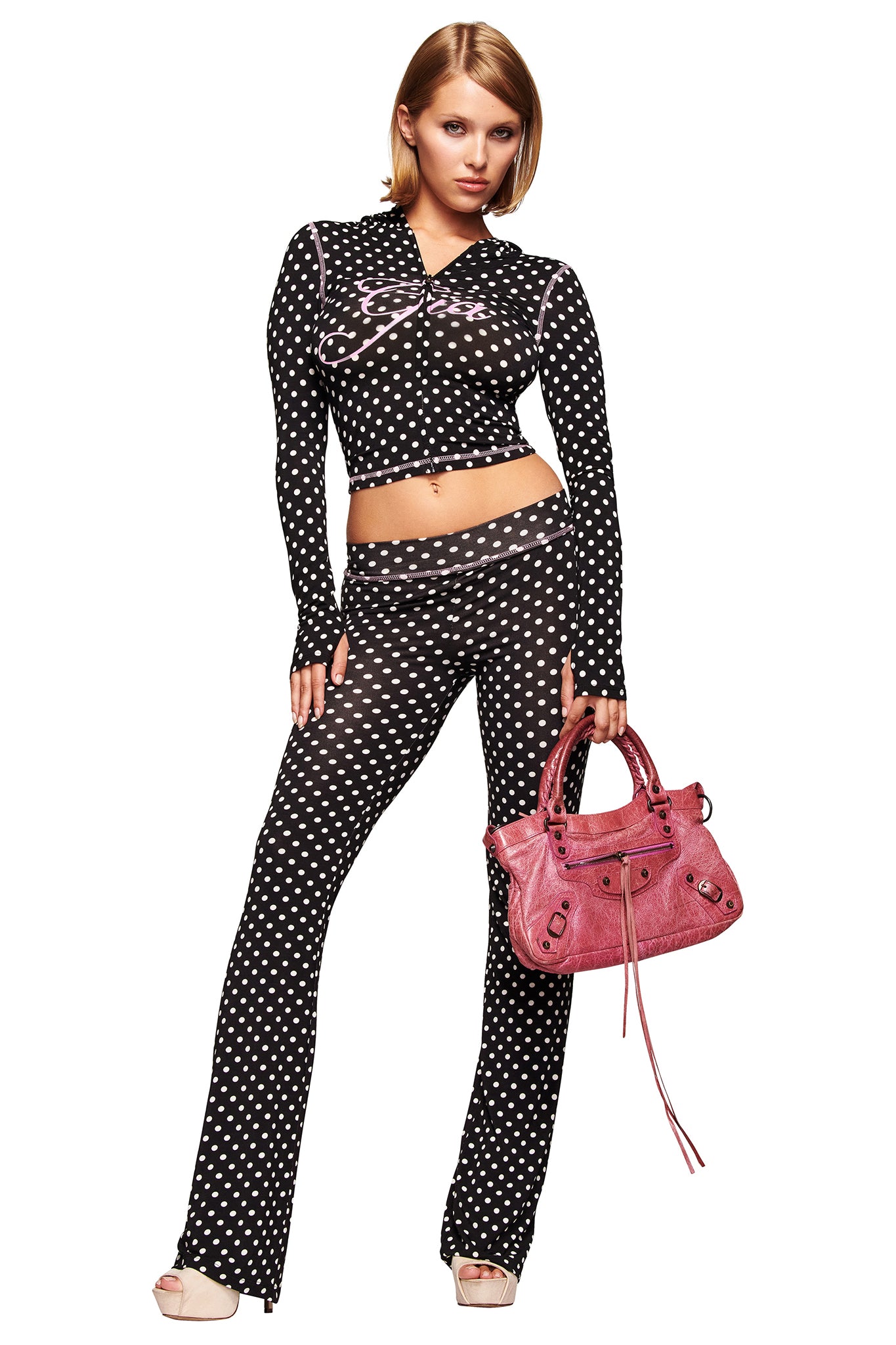 A woman with straight, shoulder-length blonde hair wears I.AM.GIA's BLARE TRACKPANT in black polka dot with a matching cropped long-sleeve top, paired with a pink handbag and light-colored platform heels.