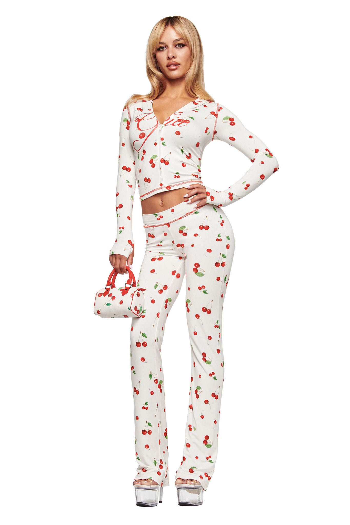 A woman with blonde hair wears the I.AM.GIA BLARE HOODIE in the Cherry on Top print, paired with matching pants and a small patterned handbag. She completes her look with silver platform heels.