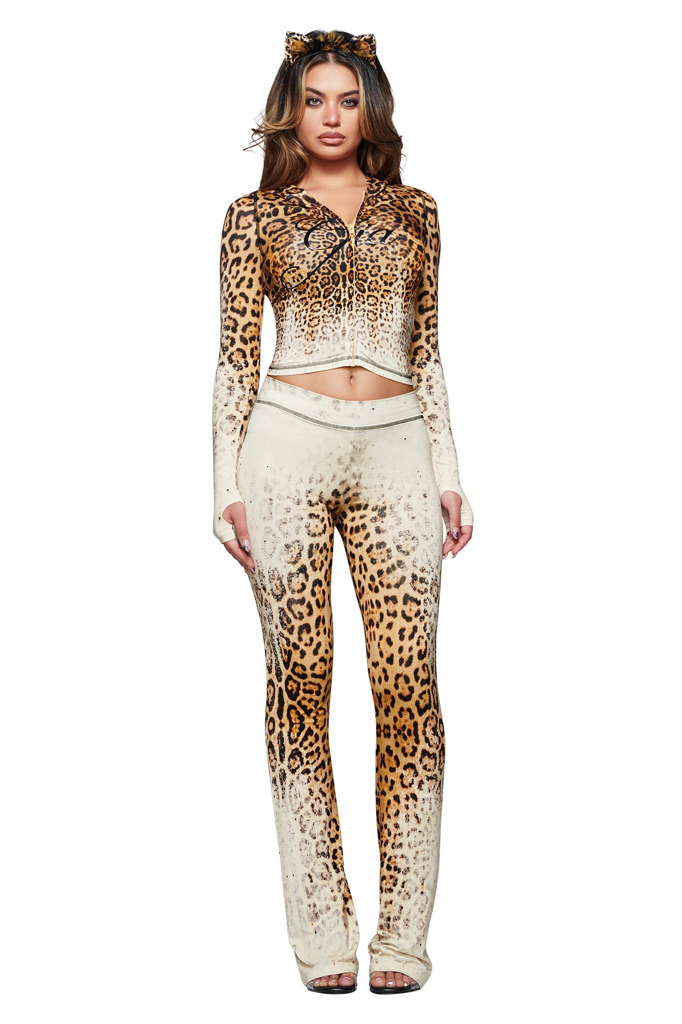 Wearing the I.AM.GIA BLARE HOODIE in Cream Faded Panthera, this woman exudes confidence as she poses against a white background. Her chic ensemble features the sustainably crafted bamboo fabrication of the matching leopard-print hoodie and pants combination, styled with thumb-hole sleeves, open-toe heels, and a coordinating leopard-print cat ears headband.