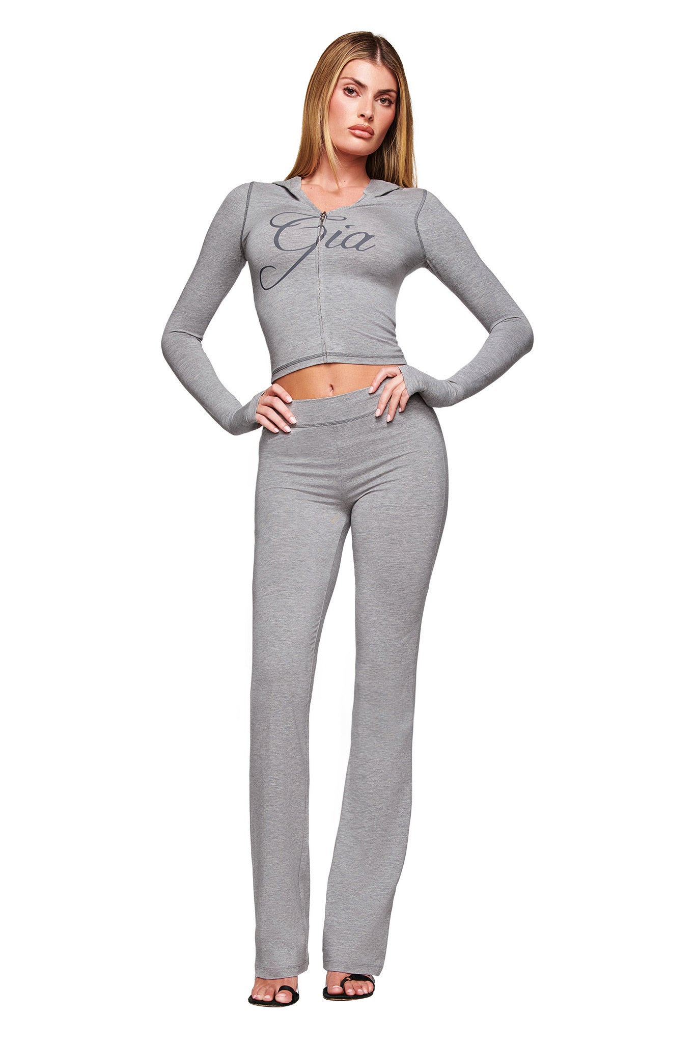 A woman faces forward in I.AM.GIA's heather grey BLARE TRACKPANT and a light gray, long-sleeve zip-up crop top with "Gia." She wears open-toe heels, has straight hair, and the flared pants stand out against a white background.