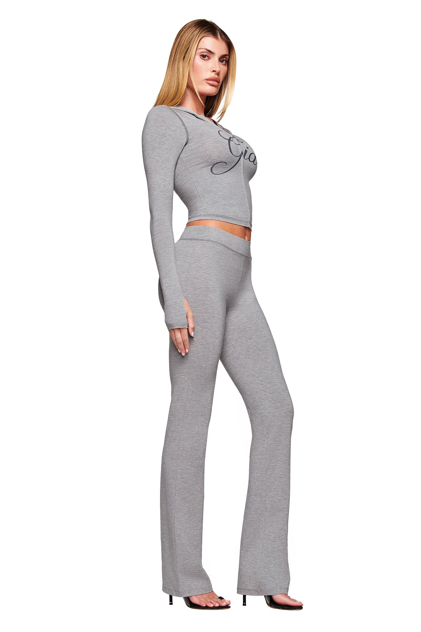 A woman poses sideways in a fitted I.AM.GIA BLARE JERSEY HOODIE - GREY: HEATHER GREY, styled as a crop top with matching high-waisted flared pants and heeled sandals against a plain white background.