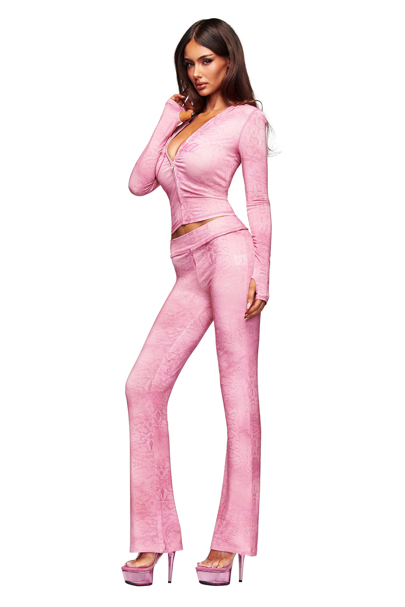 Woman poses in a light pink I.AM.GIA BLARE TRACKPANT - LIGHT PINK SERPENTINE set with long sleeves and flared pants, styled with matching platform heels, against a white background.