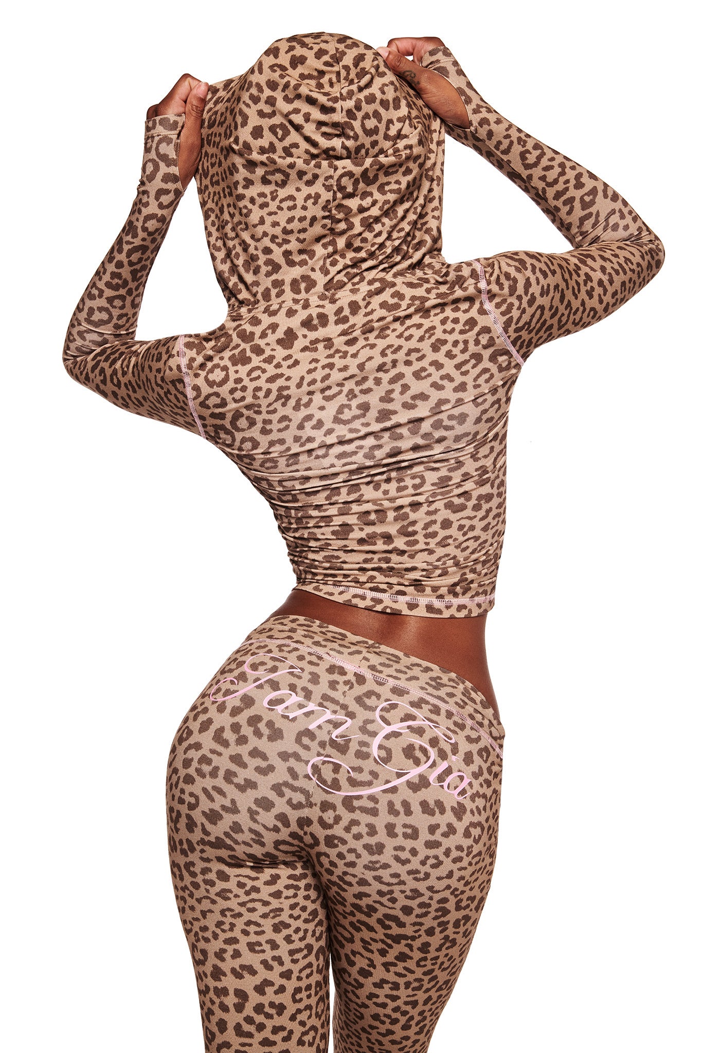 Someone in I.AM.GIA's BLARE TRACKPANT - MOCHA LEOPARD and a matching hooded top stands facing away with the hood up. The pants show cursive writing on the back, set against a plain white background.