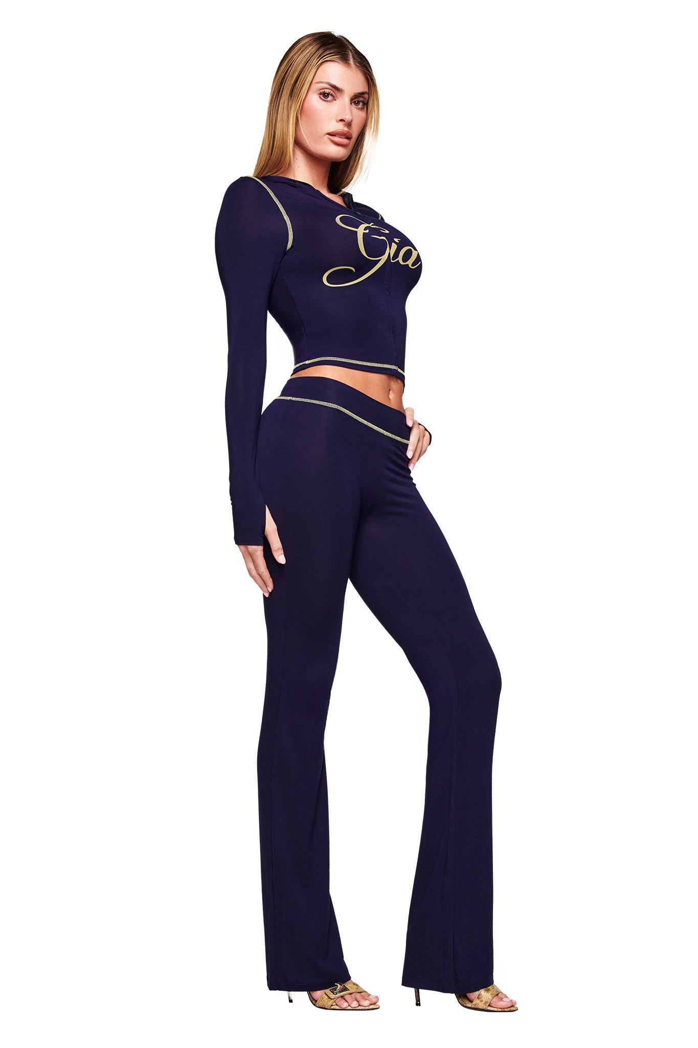 A woman poses confidently in the I.AM.GIA BLARE HOODIE - NAVY with gold script and matching bodycon, high-waisted flared pants, styled with strappy heels against a plain white background.