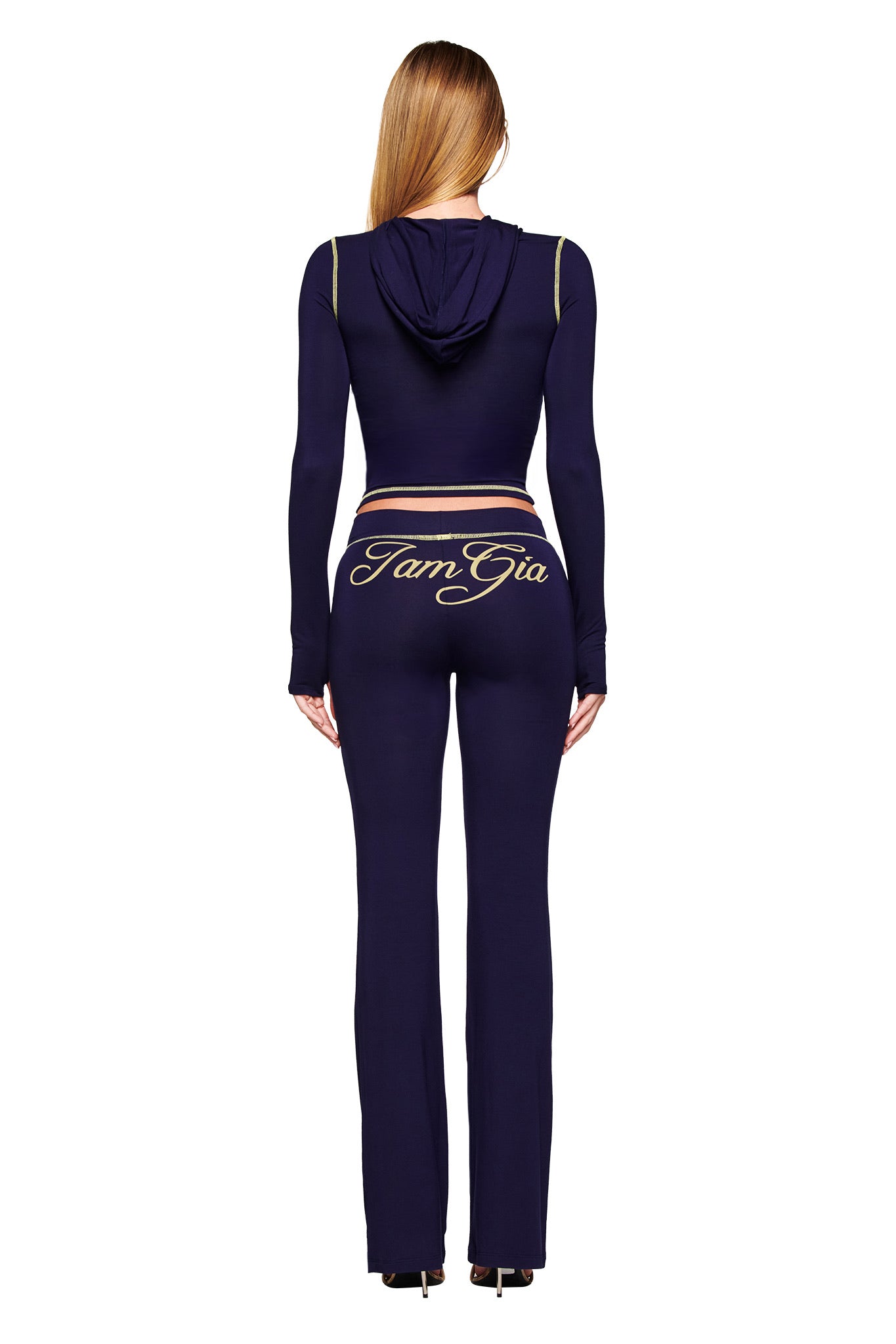 A person with long blonde hair, facing away, wears the I.AM.GIA BLARE HOODIE in navy and matching bodycon pants with gold trim. "IamGia" is printed in bold gold script on the back of the pants.